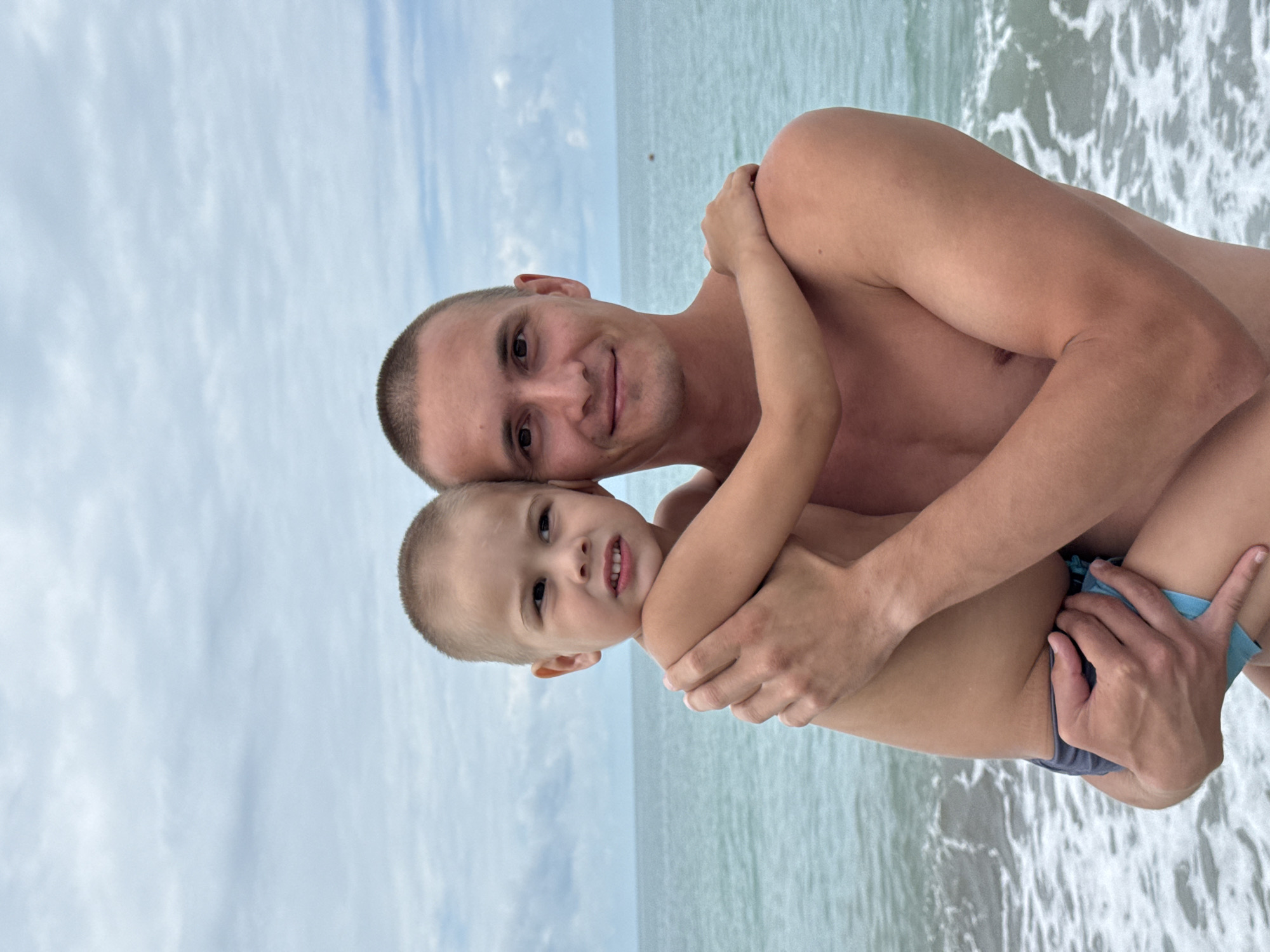 This media file captures a heartwarming moment between a father and his young son at the beach in Nebug, Russia. The father, a fair-skinned man with a shaved head and a gentle smile, is holding his son close. The son, with light brown hair and bright eyes, is nestled in his father's arms, also smiling. Both are shirtless, suggesting a warm day. The son is wearing blue swim trunks.
The background reveals the serene setting of the Black Sea. The water is a tranquil blue, with gentle waves lapping at the shore, creating a foamy white edge. The sky above is overcast with soft, diffused clouds, indicating a pleasant, possibly cooler, part of the day. The overall mood is one of love, connection, and shared enjoyment of a beach outing. No visible text is present in the media.