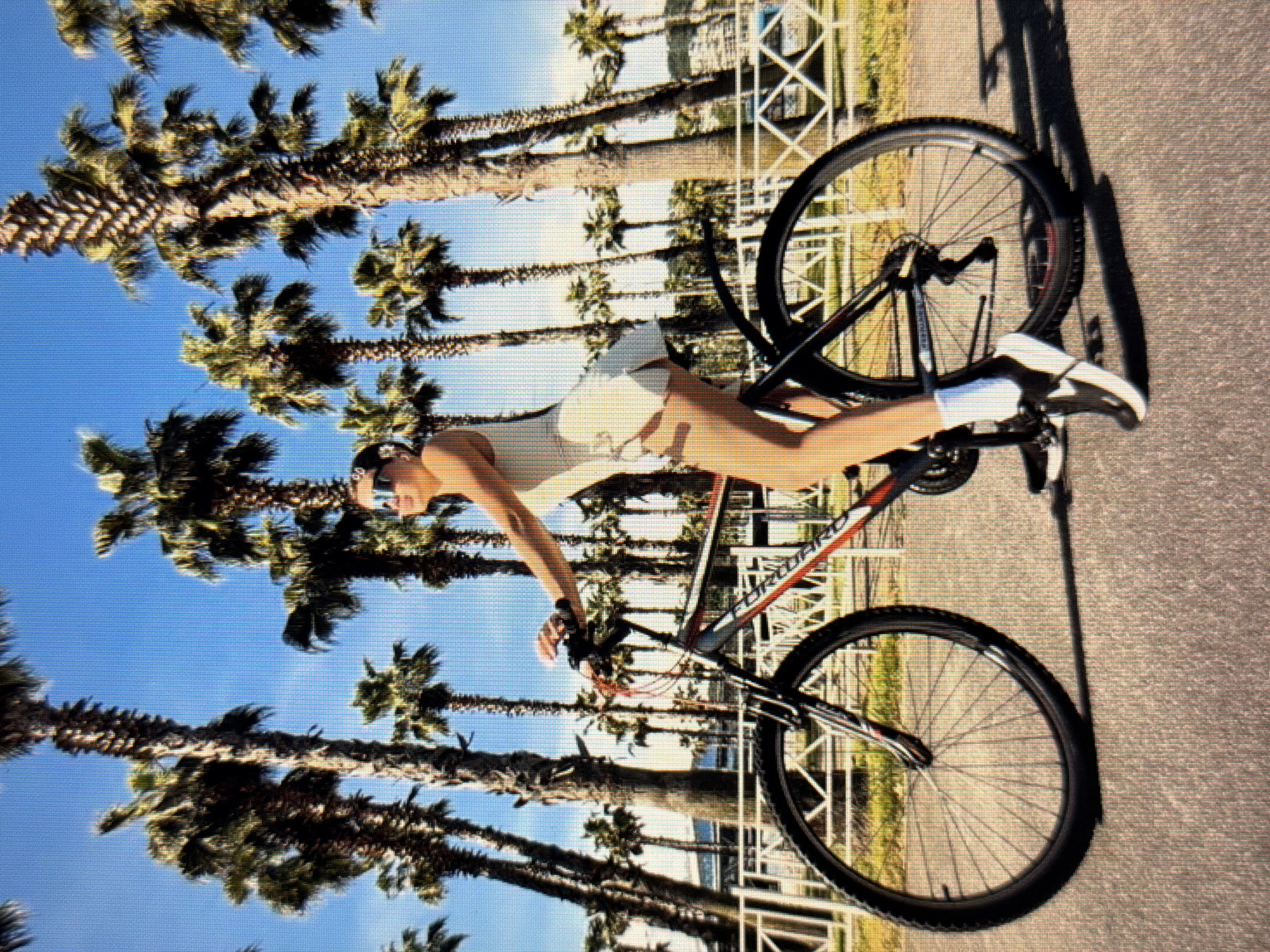 A person is riding a bicycle along a paved path lined with palm trees. The person is wearing a white athletic outfit, sunglasses, and athletic shoes. The bicycle is a dark color with red accents. The background features a clear blue sky, lush green palm trees, and a white fence. Buildings are visible in the distance behind the fence. The overall scene suggests a warm, sunny day in a coastal or tropical environment. This location is not Sirius, Russia, which is in a temperate climate and not known for palm trees.