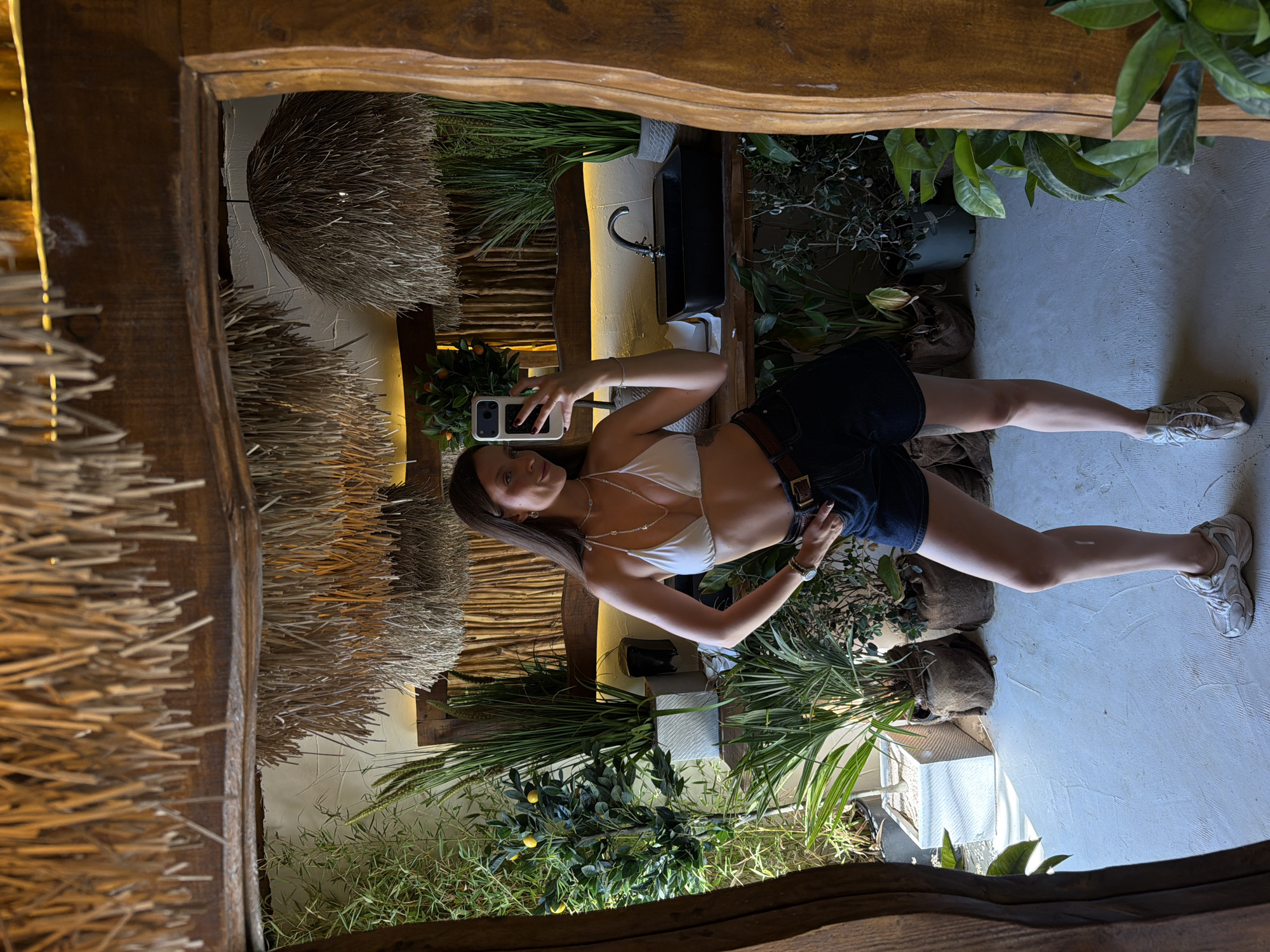A young woman with long brown hair and fair skin takes a mirror selfie in an indoor setting. She is wearing a white bikini top, dark denim shorts, a brown belt, and athletic shoes. Her left hand is on her hip, and her right hand holds a smartphone. The background appears to be a vanity area with a sink and faucet, surrounded by numerous potted plants and decorative elements, including thatched ceiling fixtures and a large wooden-framed mirror. The floor is a light-colored, smooth surface. The location is identified as Drobnići, Montenegro.