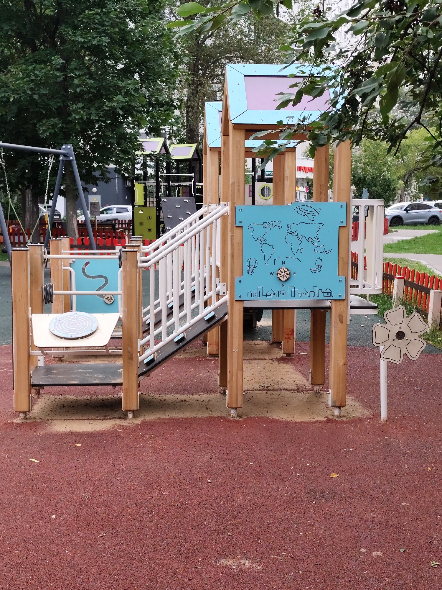 This image shows a modern children's playground in Moscow, Russia. The playground features a wooden structure with a slide, climbing elements, and interactive panels. One panel depicts a world map with small illustrations of an airplane, hot air balloon, sailboat, and a cityscape. Another panel has a maze game. A swing set is partially visible in the background, along with a red and white striped fence. The ground surface is a red rubberized material, common in modern playgrounds for safety. Trees and apartment buildings can be seen in the background, indicating an urban setting. Several cars are parked on a street adjacent to the playground. The overall scene suggests a well-maintained outdoor public space designed for children's recreation.