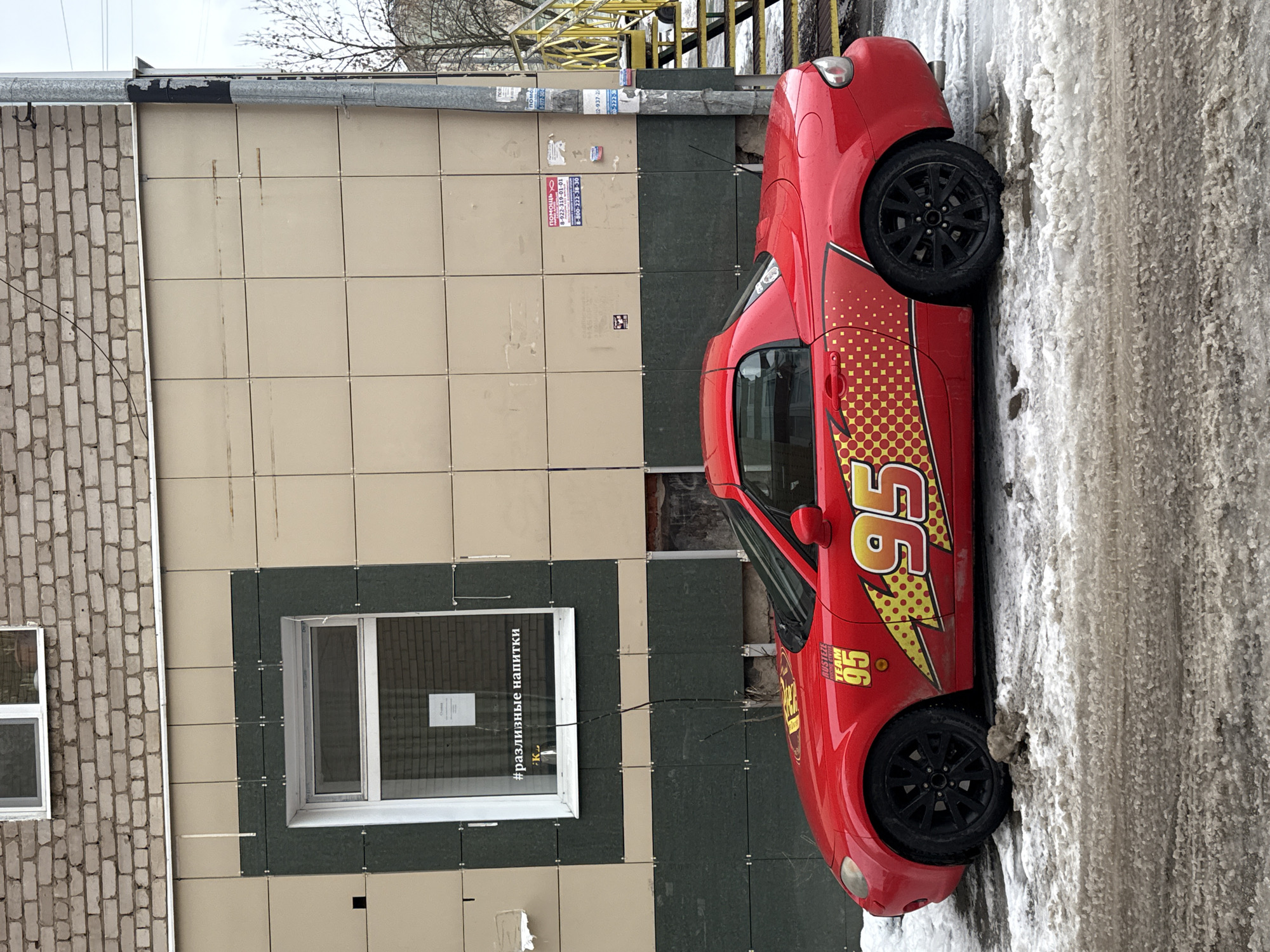 A red Mazda Miata, customized with "Cars" movie Lightning McQueen decals and the number "95", is parked on a slushy street next to a building. The building has a mix of light-colored tiles and dark green vertical panels, and a window with a sign in Russian. Debris fills gaps in the building's exterior near the car. A set of yellow metal stairs is partially visible in the background. The location is likely Perm, Russia, as indicated by the Russian text.