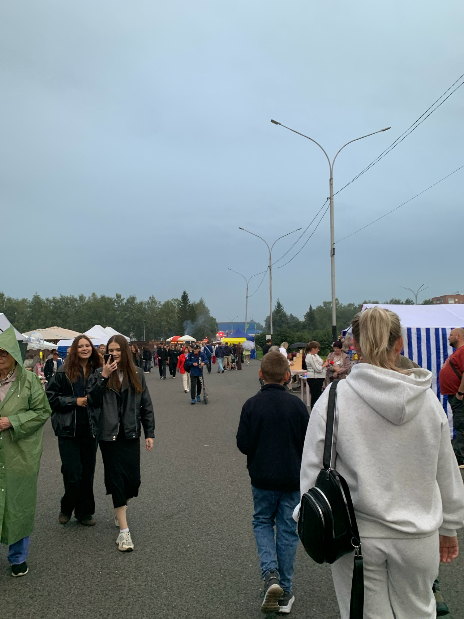 This is a vertical, eye-level, medium shot of a street market in Prokopyevsk, Russia. The sky is overcast. In the foreground, a woman with blonde hair pulled into a ponytail walks away from the camera, wearing a grey hooded sweatshirt and sweatpants. She has a black backpack slung over her left shoulder. To her left, a young boy walks away from the camera, wearing a dark blue jacket and jeans. To the left of the boy, two young women walk towards the camera, both wearing black jackets. The woman on the right is looking at her phone. To the left of the women, an elderly person wearing a green raincoat walks towards the camera. In the background, tents and stalls are set up along the street, suggesting a market or fair. Several people are visible browsing at the stalls. Streetlights and power lines are visible in the sky. The overall scene depicts a public gathering under a cloudy sky.