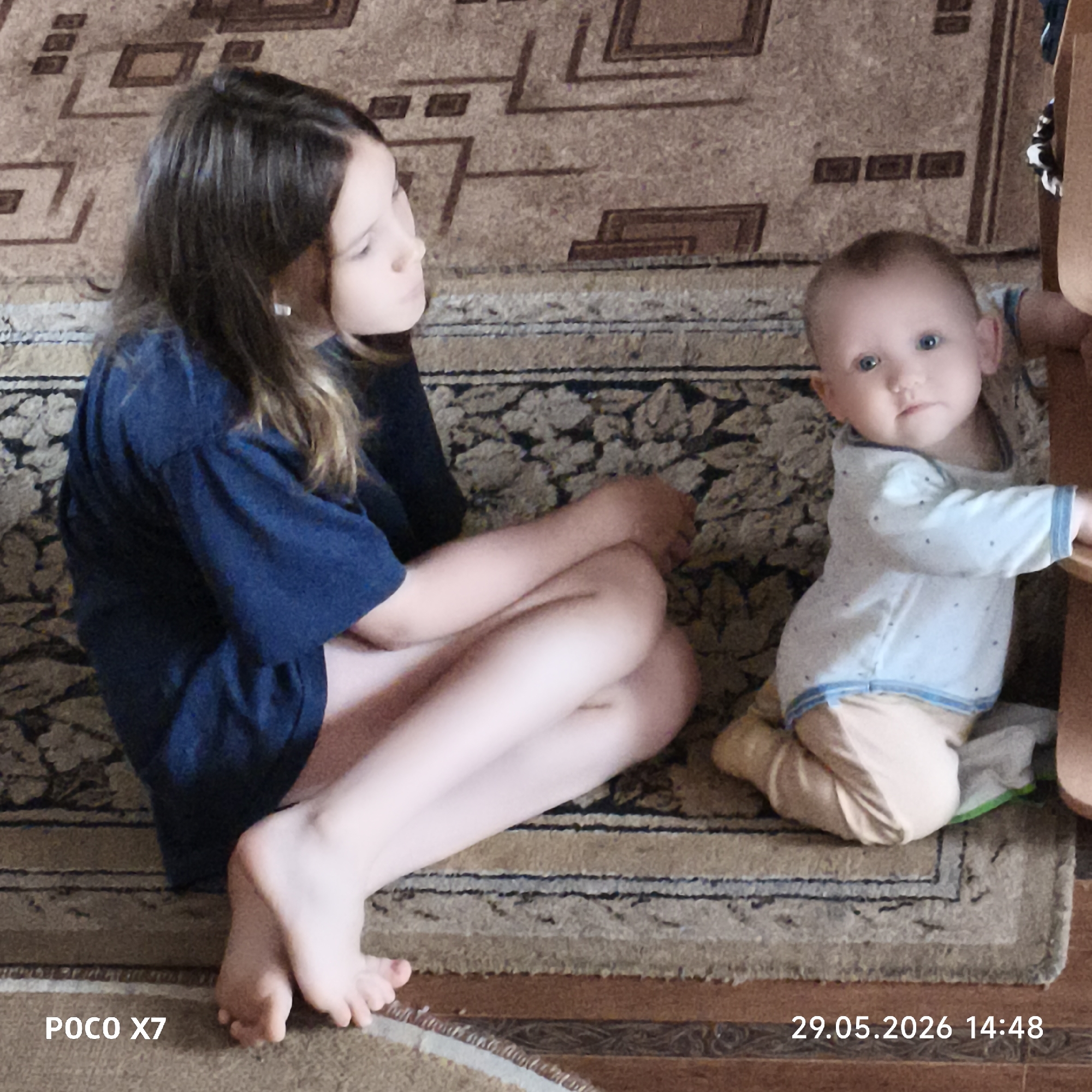 A girl and a baby sit on a carpet. The girl has brown hair and is wearing a blue t-shirt. She is sitting cross-legged and looking to the right. The baby is wearing a light blue and white long-sleeved shirt and tan pants. The baby is leaning against a wooden chair. The carpet is patterned with brown and beige designs. The photo was taken on May 29, 2026 at 14:48 with a POCO X7. The location is Unknown, Unknown.