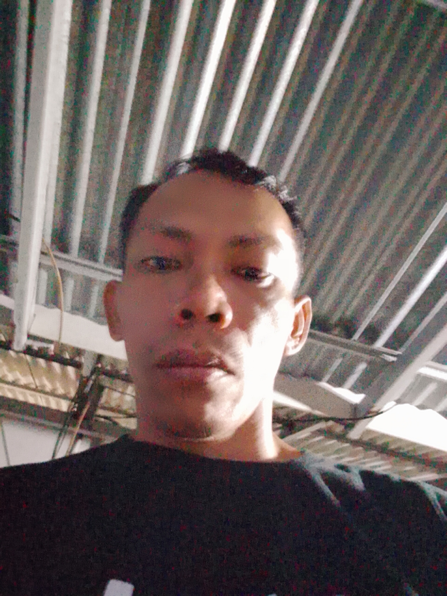 A person with dark hair and brown skin is in the foreground. They are wearing a black shirt with a white design on the bottom. The background shows a corrugated metal ceiling and exposed wiring. The location is Bukit Ketok, Indonesia.