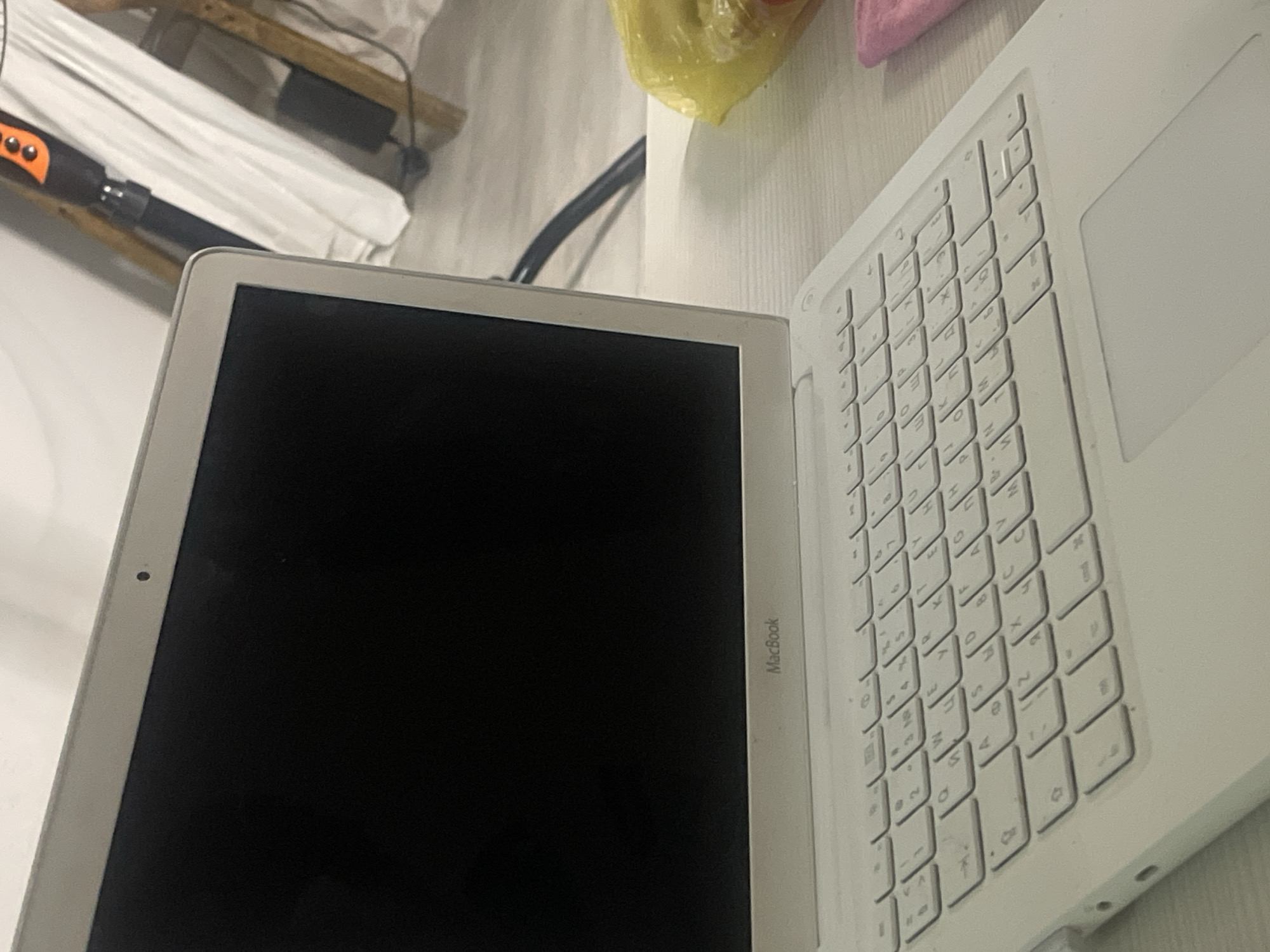 This media file captures a close-up view of an open white MacBook laptop placed on a light-colored, textured surface, likely a desk or table. The laptop is angled, showcasing its keyboard and a portion of its screen. The keyboard is primarily white with light gray lettering. To the left of the keyboard, the edge of the black screen is visible. The "MacBook" text is subtly imprinted on the bezel below the screen.
In the background, out of focus, there are hints of the setting. On the left side, a white cloth or sheet is draped over an object, and partially visible is an orange and black electronic device, possibly a tool or a charger. Further back, a black cable or power adapter is present. In the upper right corner, a yellow plastic bag and a pink object are visible, suggesting personal items.
The lighting appears to be ambient, possibly from an indoor source, casting soft shadows. The overall impression is that of a personal workspace. No people are visible in the frame. The image composition focuses on the laptop and its immediate surroundings, creating an intimate perspective. The setting appears to be indoors, given the presence of electronics and indoor lighting. There are no clear indicators of the time of day or weather conditions. The location context provided is Bishkek City, Kyrgyzstan, though there are no specific visual cues within the image to confirm this location.