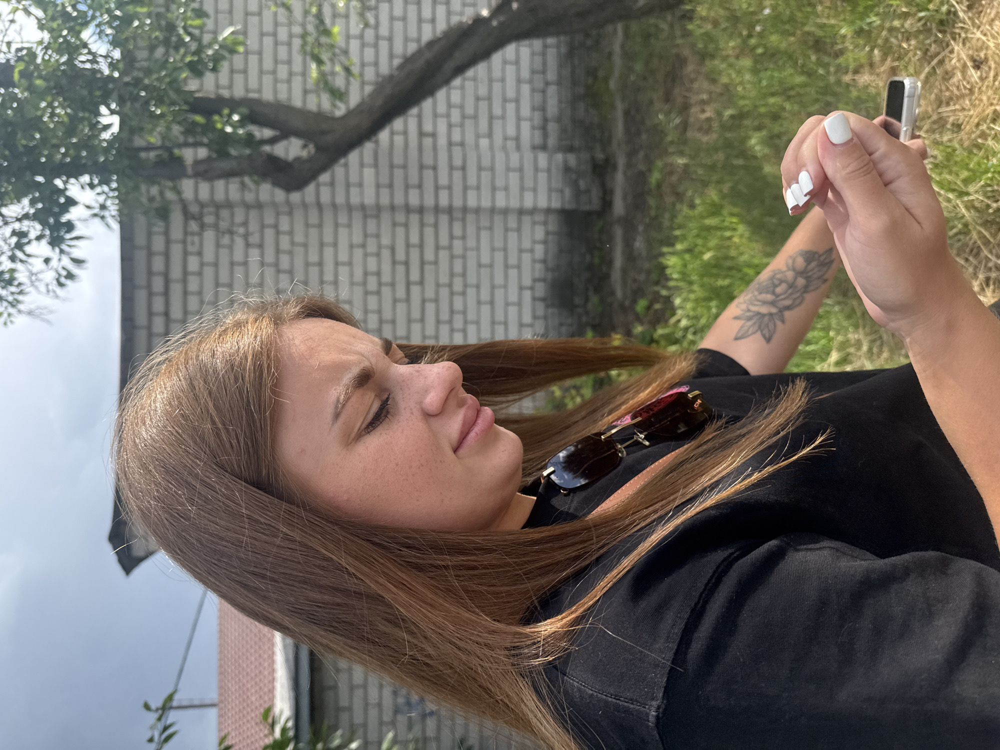 A woman with long, auburn hair, wearing a black t-shirt, is outdoors. She is looking down at her hands, which are holding a small, silver electronic device. Her fingernails are painted white. A tattoo of a flower is visible on her left arm. Sunglasses are hanging from the collar of her shirt. The background consists of a white brick wall, a tree with dark bark, and green grass. The sky is overcast. This scene is located in the city of Dnipro, Ukraine.