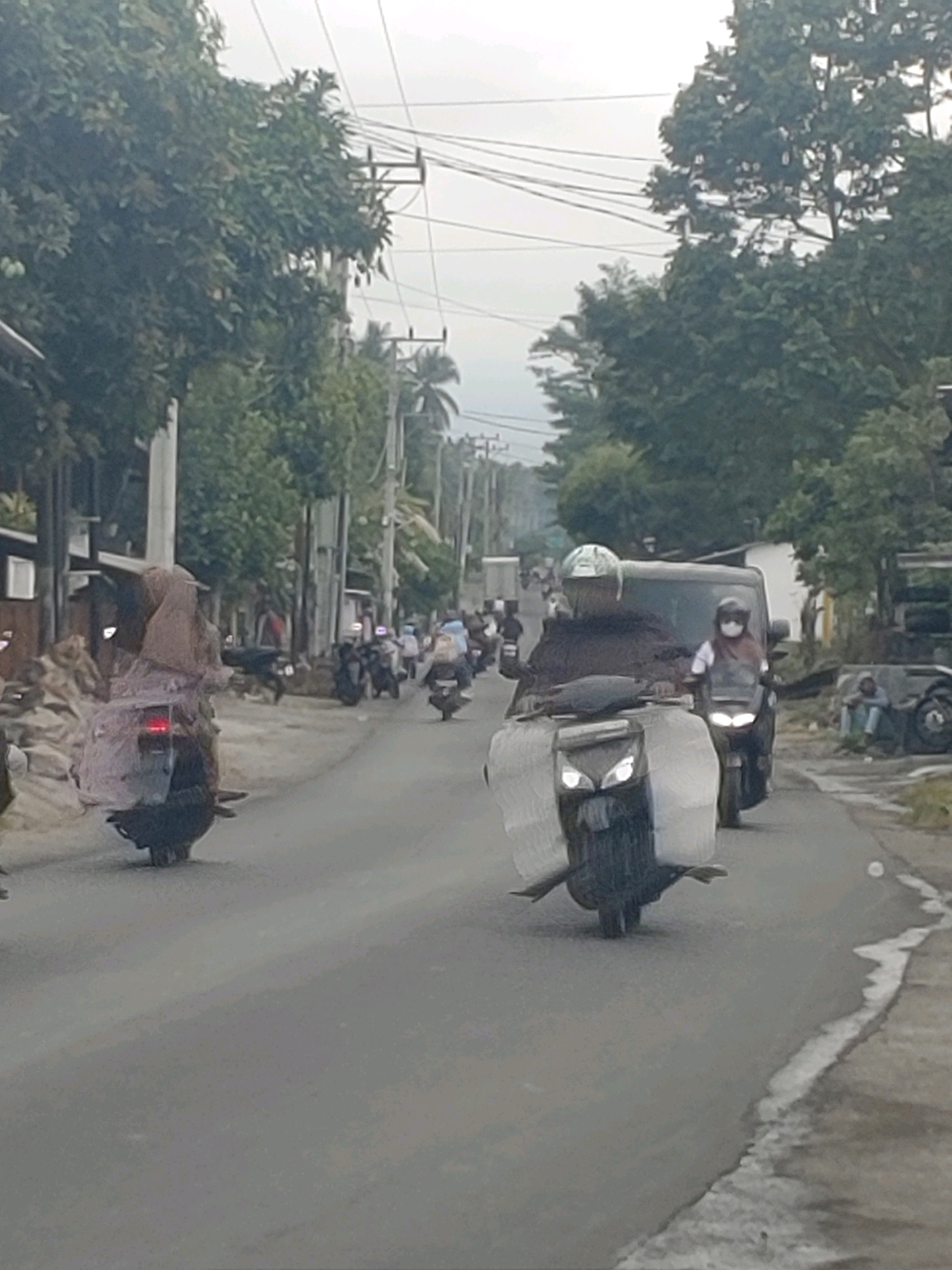 The image depicts a street scene in Kembang Kerang, Indonesia. The road is paved and appears to be a two-lane street. On the left side of the road, there are buildings with visible walls and roofs, and trees line the street. Power lines are strung overhead. The street is populated with several motorcycles. In the foreground, a person is riding a motorcycle with a large, light-colored object secured to the back. Further down the road, other motorcycles are visible, some carrying passengers. One motorcycle has a person wearing a headscarf and mask. A dark-colored van is also present on the road, with a motorcycle following behind it. On the right side of the road, there is a person sitting on the ground next to what appears to be debris or discarded materials. The overall atmosphere suggests a typical day in a town with moderate traffic.