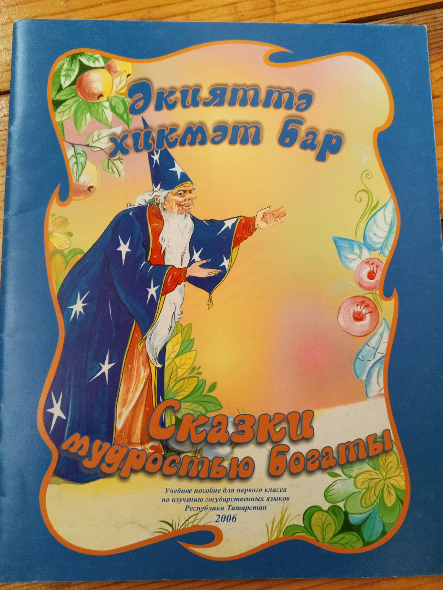 This is a close-up, eye-level shot of the cover of a children's book. The book's cover features a blue background with a decorative wavy border. Inside the border, there is an illustration of a bearded wizard in a blue, star-patterned robe and a pointed hat, gesturing with his right hand. The wizard is standing in front of a yellow and pink gradient background, with stylized green leaves and pink flowers on the right side. The top of the cover has text in a blue, stylized font, and the bottom has smaller black text. The book cover appears to be made of paper and is placed on a wooden surface. There are no discernible landmarks or elements that would indicate a specific location within the city of Chistopol, Russia.