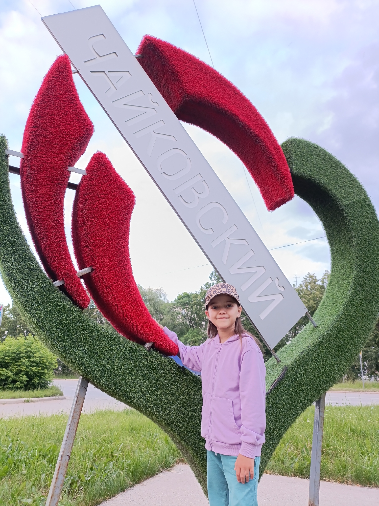 A young girl wearing a purple hooded jacket, teal pants, and a leopard-print baseball cap stands in front of a large, heart-shaped sculpture covered in green artificial grass, with two red petal-like shapes protruding from the center. A white sign with Cyrillic lettering, reading "ЧАЙКОВСКИЙ" (Chaikovsky), is diagonally positioned across the sculpture. The sculpture is located outdoors, with a street and grassy areas visible in the background, under a cloudy sky. The girl is smiling and looking at the camera, with her left hand resting on the green portion of the sculpture. The sculpture appears to be a landmark in the city of Chaikovsky, Russia, as indicated by the lettering on the sign.
