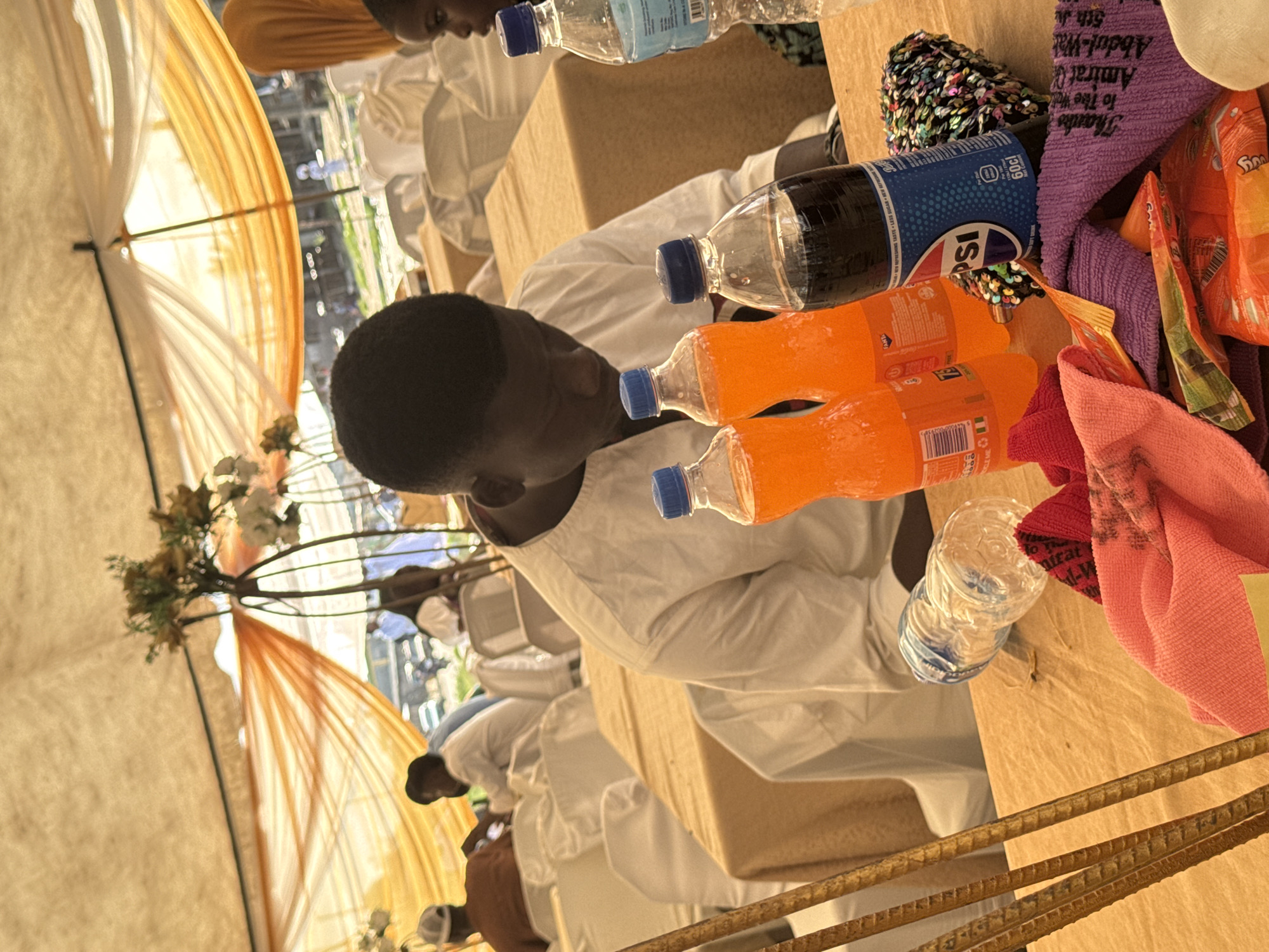 This image depicts an outdoor event likely in Badagry, Nigeria, characterized by the presence of a tented structure with draped orange and white fabric. A man in a white traditional garment is the central figure, seated and looking down at a table laden with beverages and other items. Among the visible objects are several plastic bottles of soda, including two orange-colored Fanta bottles and one bottle of Pepsi. Adjacent to these are a clear plastic bottle of water and a small, sequined object, possibly a pouch or clutch. Several colorful cloths and snack wrappers are also scattered on the table, suggesting a gathering or celebration. In the background, other individuals are visible, some seated at tables covered with beige tablecloths, and chairs draped in white fabric. The overall setting suggests a communal event, possibly a wedding reception or a similar festive occasion, taking place in an open-air environment.