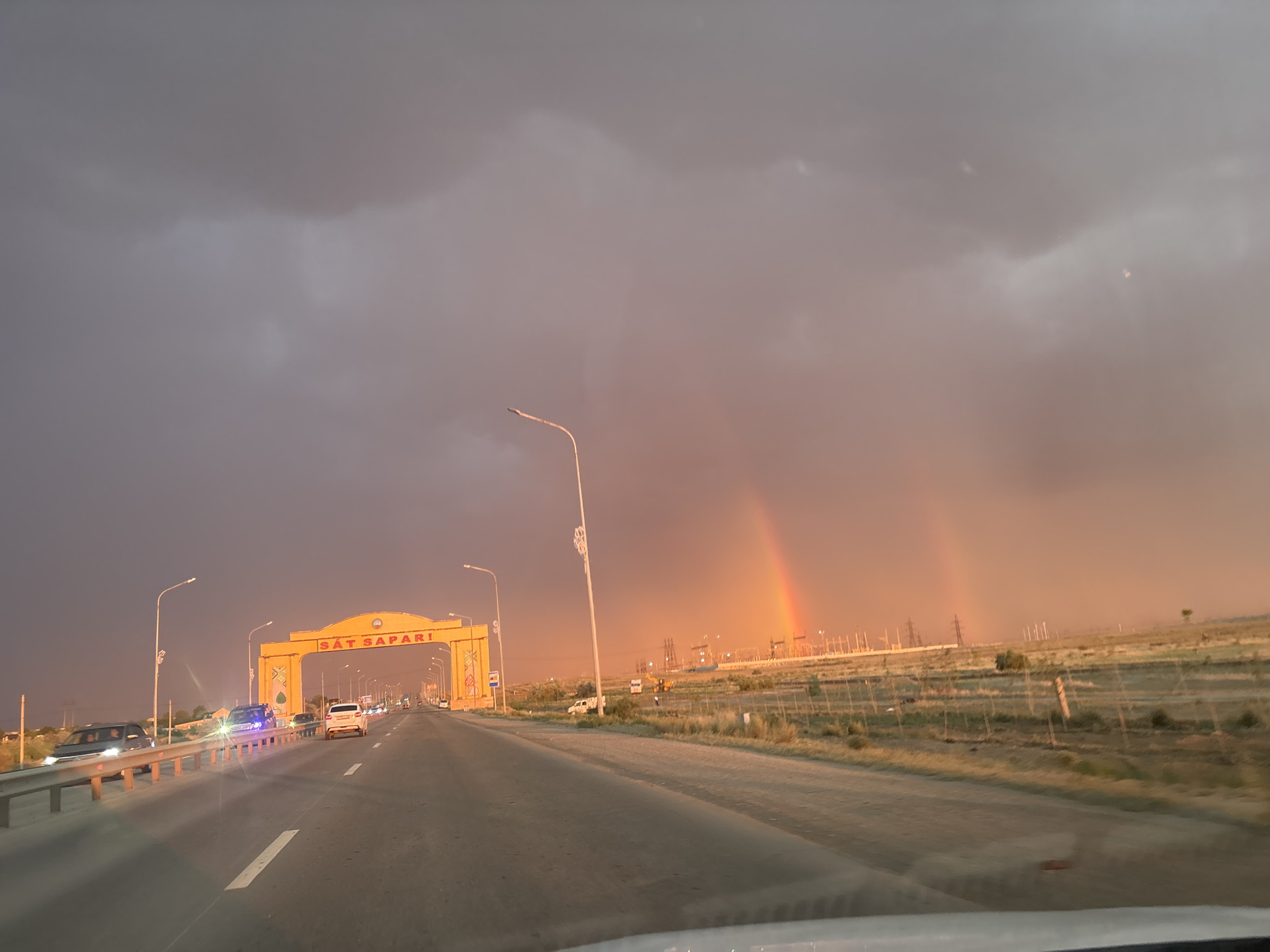 The image depicts a scenic drive under a dramatic sky, likely during a transition between day and evening. The scene is set on a highway in Satbayev, Kazakhstan, with a prominent yellow archway spanning the road bearing the text "SAT SAPARI" in red letters. This archway serves as an entrance or landmark.
The sky is filled with dark, ominous clouds, suggesting recent or impending rain. However, a beautiful double rainbow has emerged, arcing across the sky and casting a warm, colorful glow on the horizon. This juxtaposition of stormy clouds and a vibrant rainbow creates a visually striking and somewhat emotional atmosphere, perhaps evoking feelings of hope or awe.
Several vehicles are visible on the highway, including cars and what appears to be a truck or bus, indicating regular traffic. Streetlights are also present, some of which are illuminated, suggesting that the time of day is either early morning or late afternoon/early evening, as the natural light fades. In the background, there are power lines and what might be industrial or infrastructure structures, characteristic of a developed area. The landscape is predominantly flat and arid, with dry grass and sparse vegetation lining the sides of the road. The overall impression is of a journey through a vast landscape, marked by both the power of nature and human infrastructure.