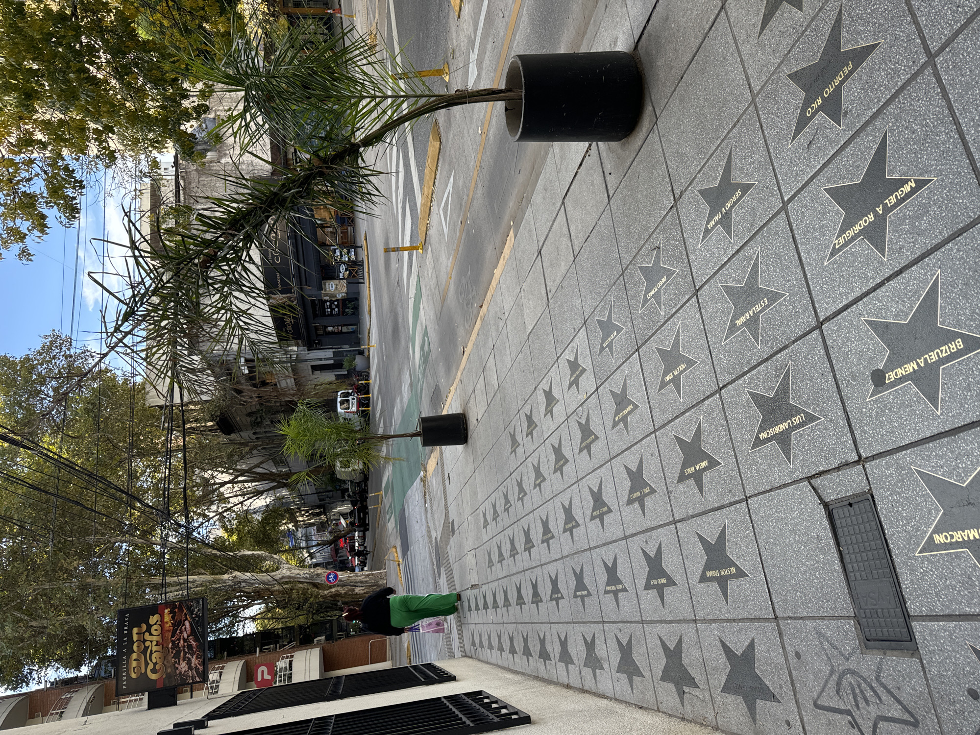 The image depicts a sidewalk in Buenos Aires, Argentina, lined with grey tiles bearing embedded star-shaped plaques. Each plaque displays a name, presumably honoring individuals. Notable names include "MARCONI", "BRIZUELA MENDEZ", and "PEDRITO RICO". A woman wearing a green skirt or pants can be seen standing on the sidewalk. Black cylindrical planters containing trees are positioned along the edge of the sidewalk. Beyond the sidewalk, the street is visible. A building with a large advertisement that says "Don Carlos" is visible in the background. Power lines crisscross above. The sky is partially visible, suggesting a sunny day.
