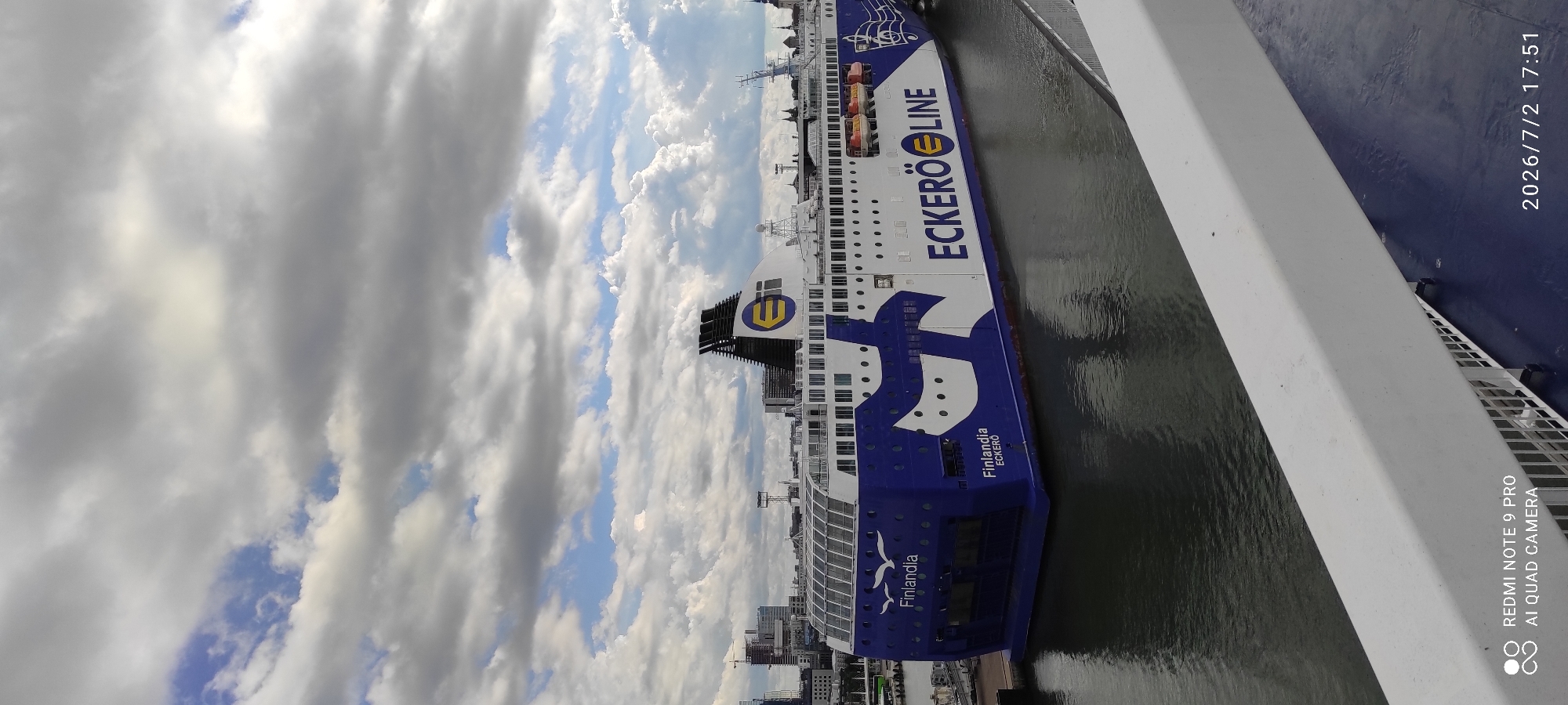 The media file shows a ferry docked in a harbor under a cloudy sky. The ferry, named "Finlandia" and operated by "ECKERÖ LINE," is predominantly blue and white with the company's logo visible. The ferry appears to be stationary, suggesting it is either about to depart or has just arrived. The water in the harbor is calm and reflects the sky and the ferry. In the background, city buildings and construction cranes can be seen, indicating an urban setting. The timestamp on the image reads "2026/7/2 17:51," suggesting the photo was taken in the late afternoon of July 2, 2026. The weather appears partly cloudy, with sunlight breaking through the clouds in some areas. The image is taken from an elevated perspective, likely from another vessel or a building overlooking the harbor. The text "REDMI NOTE 9 PRO AI QUAD CAMERA" is also visible, indicating the type of device used to capture the image.