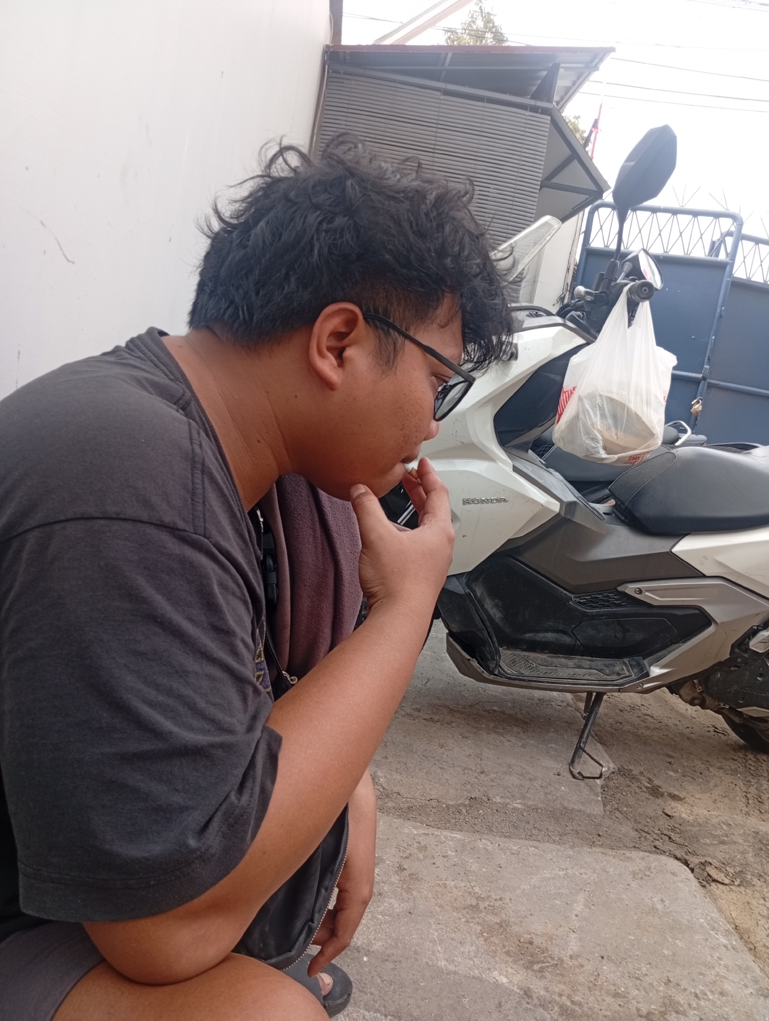 An Indonesian person with curly black hair and glasses is sitting on the ground, facing left, with their right hand near their mouth, possibly smoking. They are wearing a dark grey t-shirt and dark shorts. A light-colored plastic bag hangs from the seat of a white Honda scooter parked behind them. The scooter is parked on a cracked concrete surface. In the background, there is a white wall, a metal gate, and a corrugated metal roof structure. The environment appears to be outdoors, possibly in a residential or urban area.