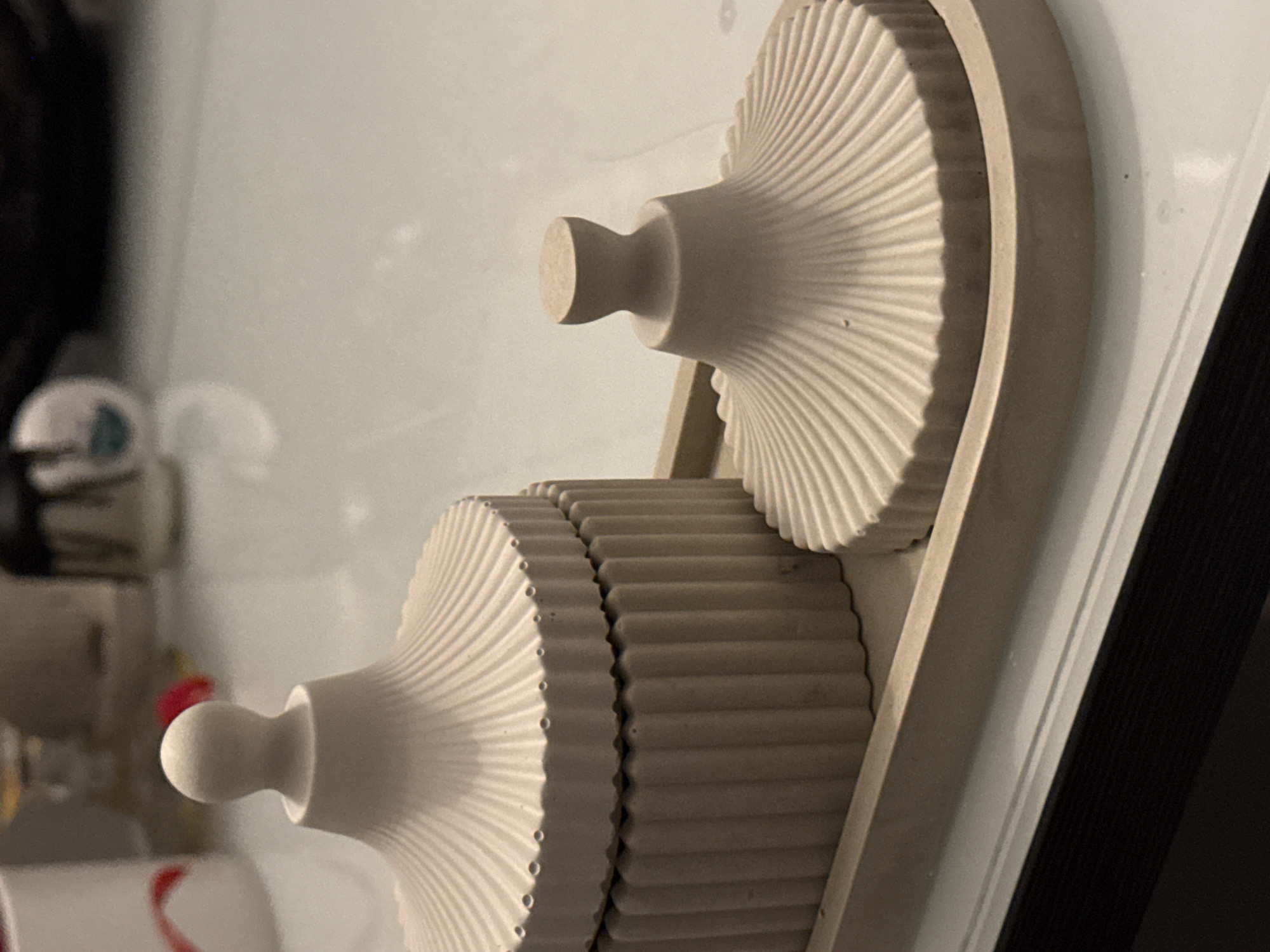 A close-up shot shows two bisque-colored cylindrical gears with prominent radial fluting and bulbous handles, mounted on an off-white frame attached to what appears to be a white surface. The top gear is a single cylinder, while the lower gear consists of two cylinders side-by-side. A black vertical edge is visible on the right side. A blurry background suggests the image was taken indoors, but the exact location is indeterminable.