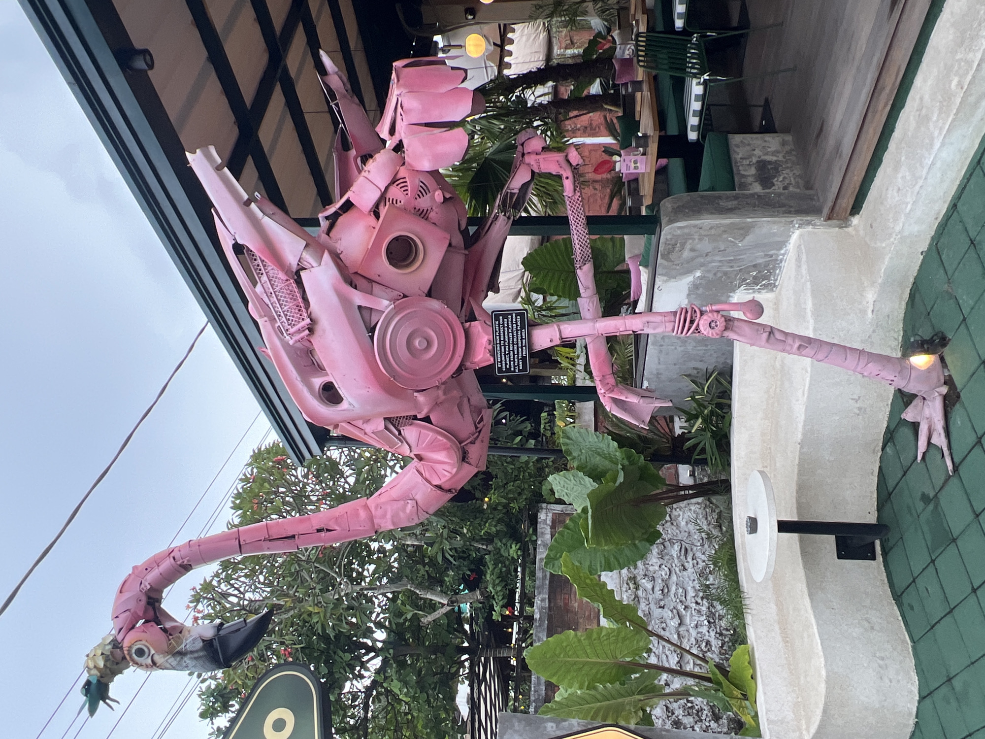 A large, vibrant pink flamingo sculpture, crafted from recycled plastic and iron, stands prominently in an outdoor setting in North Kuta, Indonesia. The sculpture, resembling a robotic or mecha-flamingo, features intricate details including gears, grates, and repurposed components forming its body, long neck, and slender legs. Its head has a detailed beak and eye, with a small green element attached.
The scene appears to be at dusk or late afternoon, with soft, diffused light and some artificial lights visible in the background. The flamingo is positioned near a concrete structure, possibly a planter or wall, and surrounded by lush green foliage, including large-leafed plants and trees with small red flowers, characteristic of a tropical environment. In the background, under a dark overhang with visible beams, an outdoor dining area with green chairs and wooden tables can be discerned.
A small black plaque attached to the sculpture's body contains text emphasizing its construction from recycled materials and promoting environmental responsibility: "BY REDUCING OUR PLASTIC CONSUMPTION, THIS FLAMINGO WAS MADE FROM RECYCLED PLASTIC AND IRON, WE NEED TO MAKE SURE EVERY TRASH DISPOSAL PLACES FUND THE AREA." The overall impression is one of creative environmental art set in a relaxed, green outdoor space.