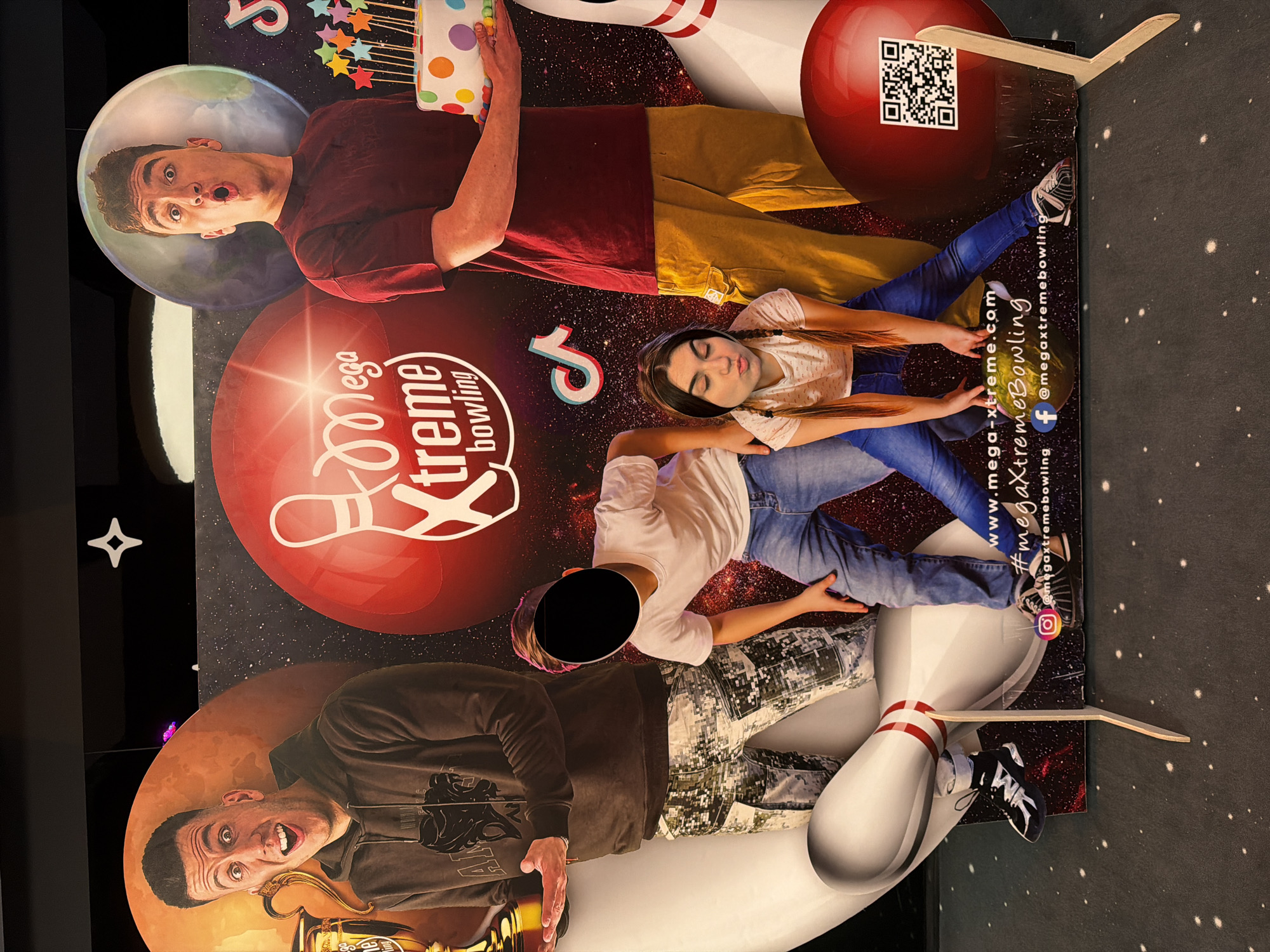A colorful advertisement is displayed, featuring a group of people superimposed over an image of bowling pins, balls, and a background of cosmic space. A man at the top left is making a surprised expression, holding a birthday cake. Underneath him is the logo "Mega Xtreme Bowling" above a girl with her eyes closed. Beneath them is another person with their face blacked out. At the bottom left, a man with his mouth open is holding a gold trophy. QR codes and social media contact information can be seen on the right of the image. The advertisement likely belongs to a Mega Xtreme Bowling alley, a company in Sofia, Bulgaria.