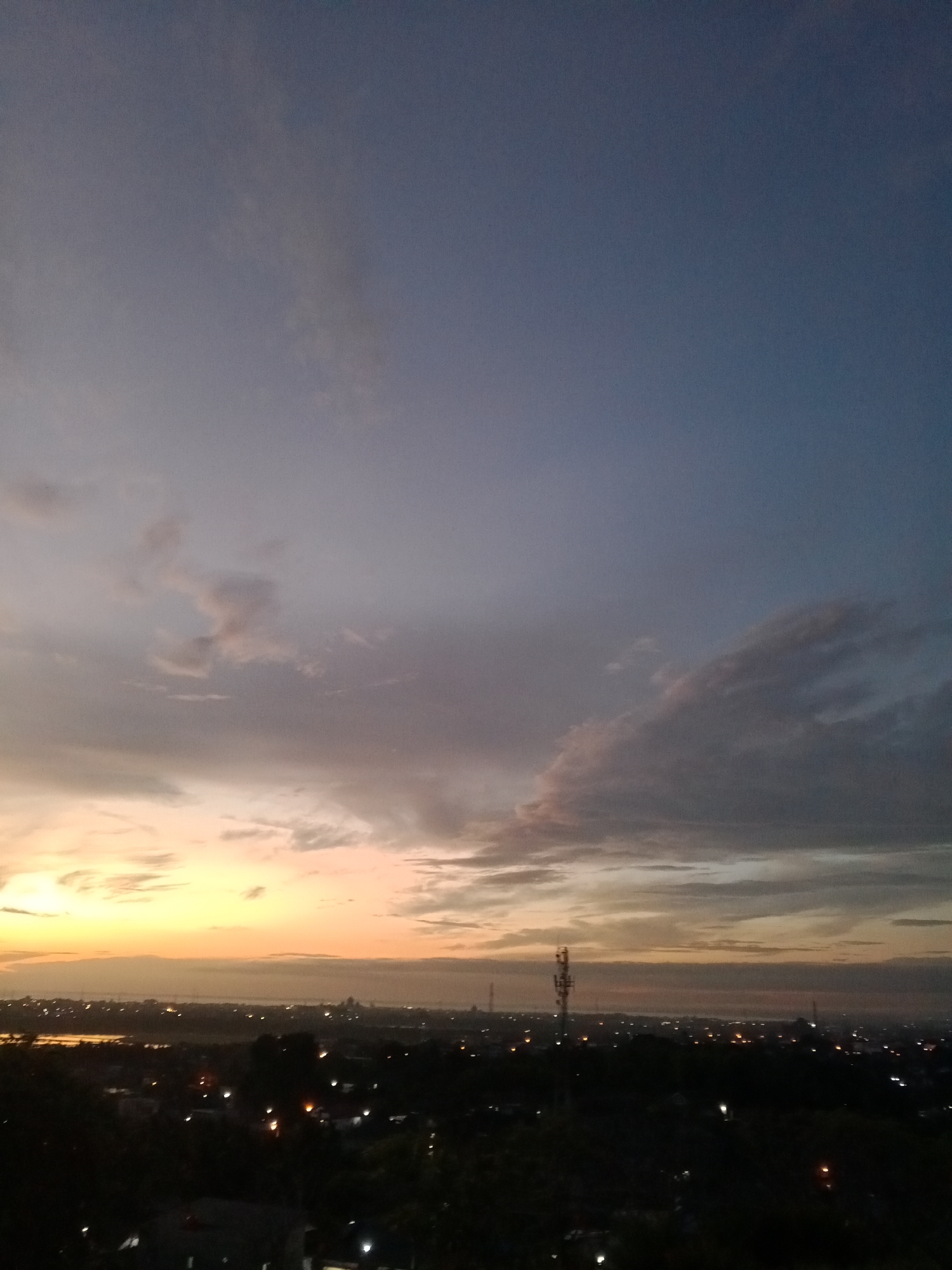 The image depicts a twilight cityscape, possibly Lhokseumawe, Indonesia, viewed from an elevated position. The sky transitions from a dark blue at the top to an orange-yellow horizon, suggesting either sunrise or sunset. Scattered clouds are present, with heavier concentrations near the horizon. Below the horizon, an urban landscape unfolds. Structures are silhouetted against the brightening sky. Lights are visible within the city. A cell tower is visible. The foreground contains darker areas of trees or built structures.