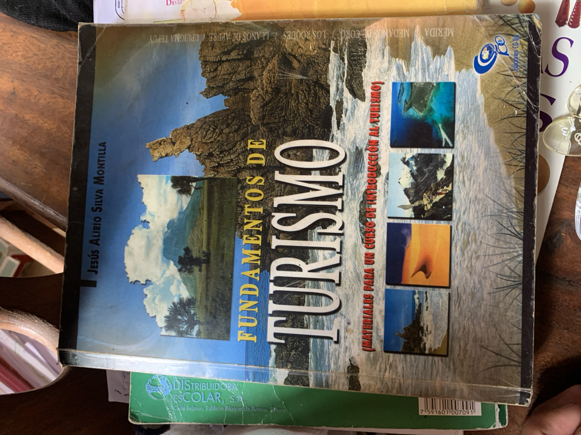 The image displays a Spanish-language textbook titled "FUNDAMENTOS DE TURISMO" (Fundamentals of Tourism) by JESÚS ALIRIO SILVA MONTILLA, set on a wooden surface, likely a table, in Santa Ana, Venezuela. The subtitle reads "(MATERIALES PARA UN CURSO DE INTRODUCCIÓN AL TURISMO)" (Materials for an Introduction to Tourism Course).
The book cover features a collage of landscape imagery. A large central image shows a dramatic rocky coastline with waves, while a scene with mountains, clouds, and palm trees is visible on the left side. Several prominent Venezuelan geographical locations are listed across the top of the cover: MÉRIDA, MÉDANOS DE CORO, LOS ROQUES, LLANOS DE APURE, and UPUIGMA TEPUY, indicating a focus on tourism within the country. To the right, four smaller square images depict various tourist-related scenes, including an underwater view, a fiery sunset over water, and another coastal area.
The publisher's logo, "Ediciones CO-BO," is visible in the upper right corner. At the bottom, a green book is partially visible underneath, bearing the name "DISTRIBUIDORA ESCOLAR, S.A." and a barcode with the number "7 591601 007091." The book shows signs of use, with some wear and tear along the edges and spine. The lighting is ambient, and no specific activity or people are depicted beyond the book itself.
