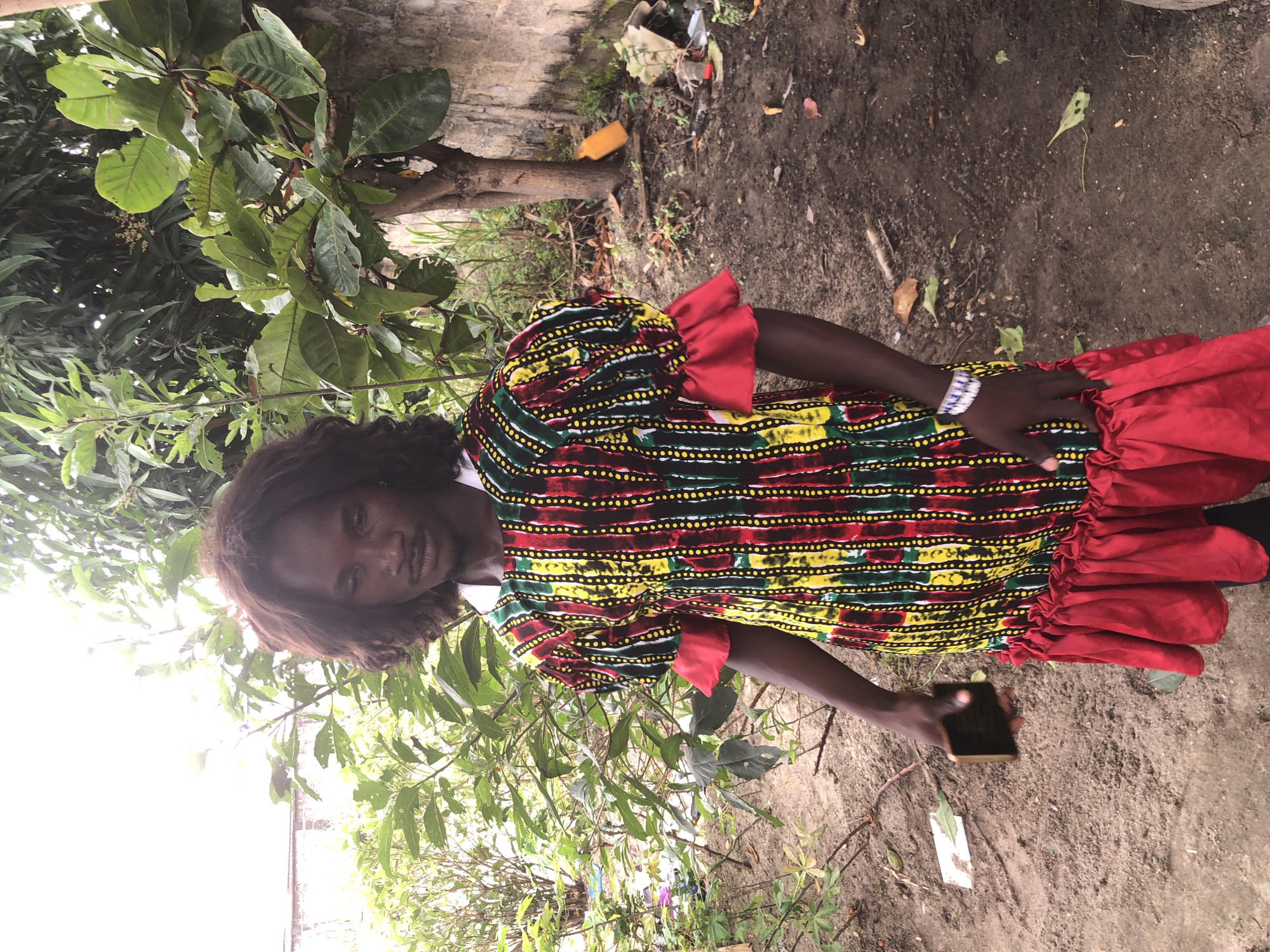A man with dark, curly hair and a mustache stands outdoors in Ado, Nigeria, holding a smartphone in his left hand. He is wearing a vibrantly colored, multi-patterned dress with ruffled red sleeves and a ruffled red hem. The dress features vertical stripes of red, green, yellow, and black, interspersed with rows of yellow dots. He also wears a beaded bracelet on his left wrist. The background consists of lush green foliage and trees, a textured concrete wall, and a dirt ground littered with debris, including plastic waste. The scene suggests an outdoor, possibly unkempt, environment in a Nigerian city.
