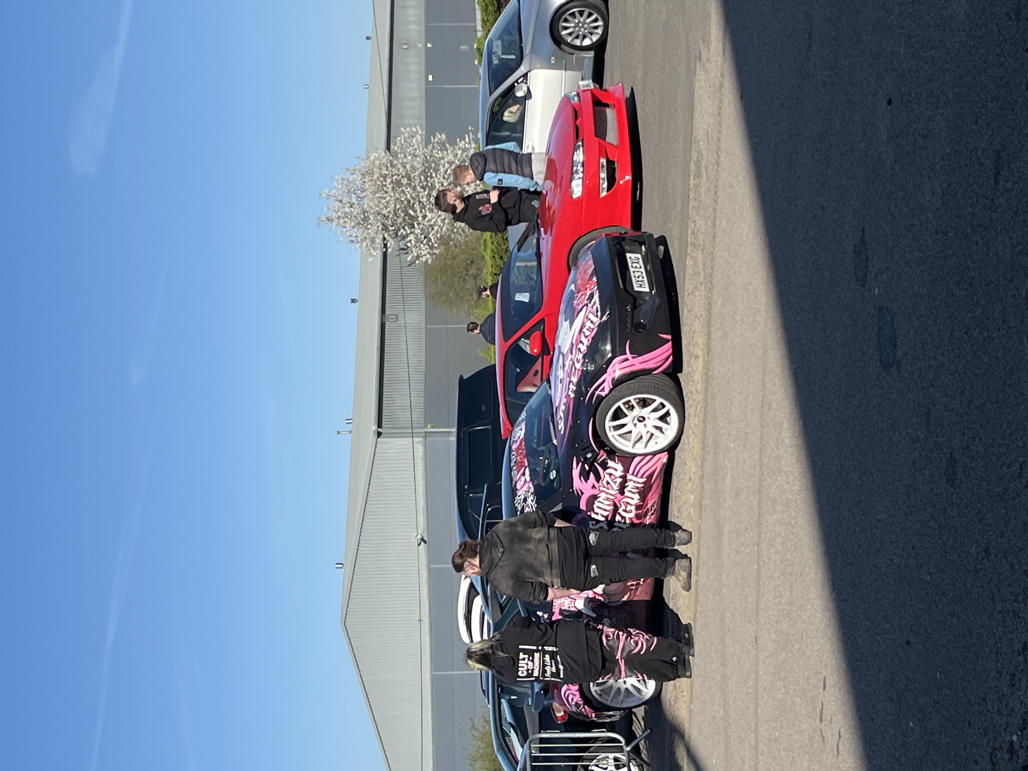 The image shows a gathering of cars, predominantly sports cars, in what appears to be a parking area. Two individuals are standing near a black car with pink decals and white wheels, one appearing to be examining the vehicle. Another two individuals are looking at a red car behind the first car. The cars are arranged in a line along a road in front of an industrial or commercial building. The location appears to be in the city of Silverstone, United Kingdom. The weather is sunny, with a clear blue sky.