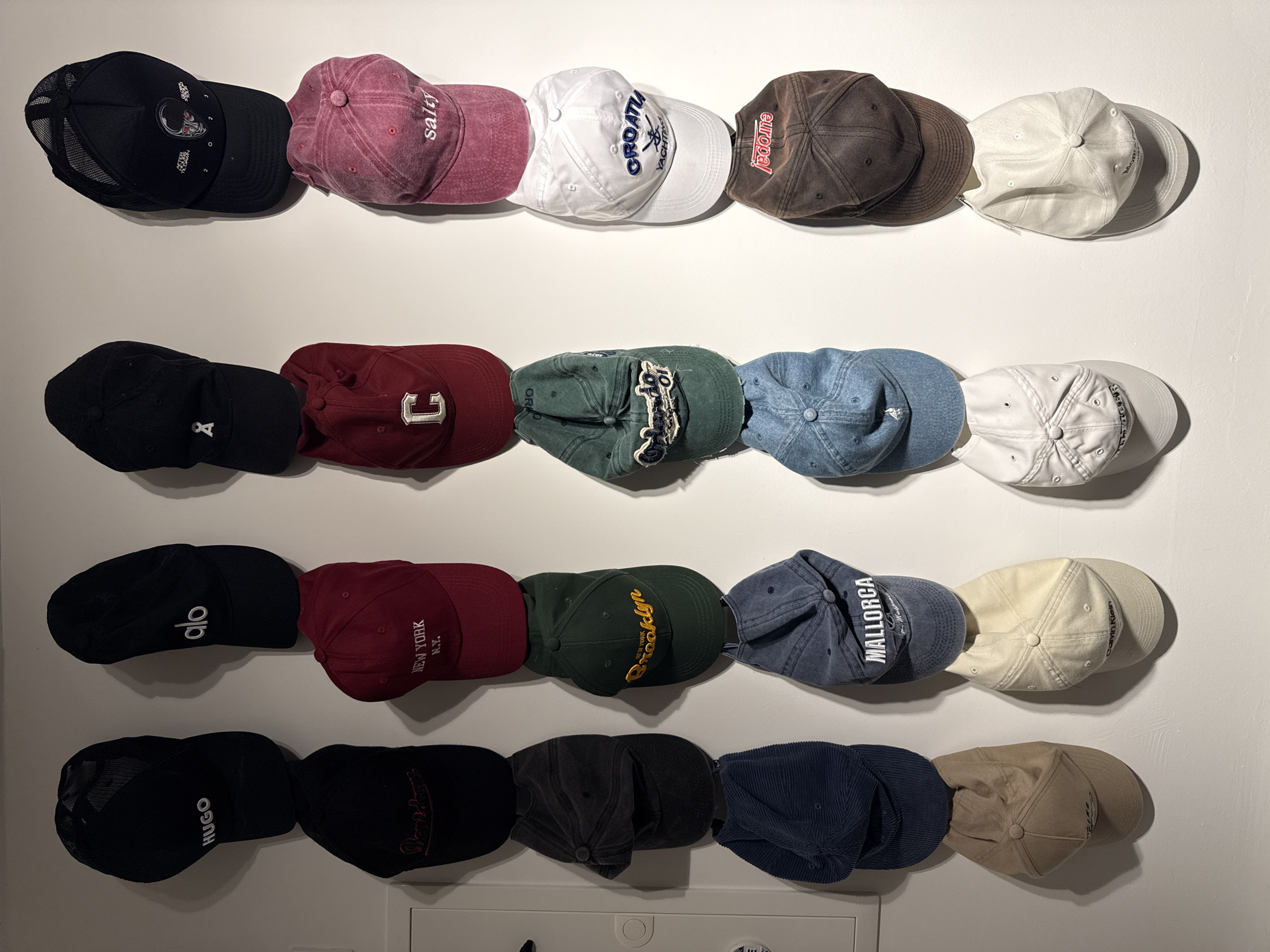 The image shows a collection of baseball caps hanging on a white wall. The caps are arranged in four vertical columns, with each column containing four caps. The caps vary in color and design, with some featuring text and logos.
**Top row, left to right:**
* Black cap with "HUGO" in white text.
* Black cap with "alo" in white text.
* Black cap with a white "x" logo.
* Black mesh trucker hat with graphics.
**Second row, left to right:**
* Black cap with red text.
* Red cap with "NEW YORK N.Y." in white text.
* Red cap with a white "C" logo.
* Pink cap with "salty" in white text.
**Third row, left to right:**
* Dark grey cap with lighter grey text.
* Green cap with "Brooklyn" in yellow text.
* Blue cap with "CROATIA YACHT CLUB" and a graphic.
* Brown cap with red text.
**Fourth row, left to right:**
* Dark blue ribbed cap.
* Blue cap with "MALLORCA" in white text.
* Light blue cap with a white logo.
* White cap with a black and red graphic.
**Bottom row, left to right:**
* Tan cap with no visible text or logo.
* Off-white cap with "Calvin Klein" in black text.
* White cap with no visible text or logo.
* Off-white cap with black text.
There are no people, actions, or interactions depicted in the image. The environment is a plain white wall, suggesting an indoor setting. The location is not specified within the image itself.
