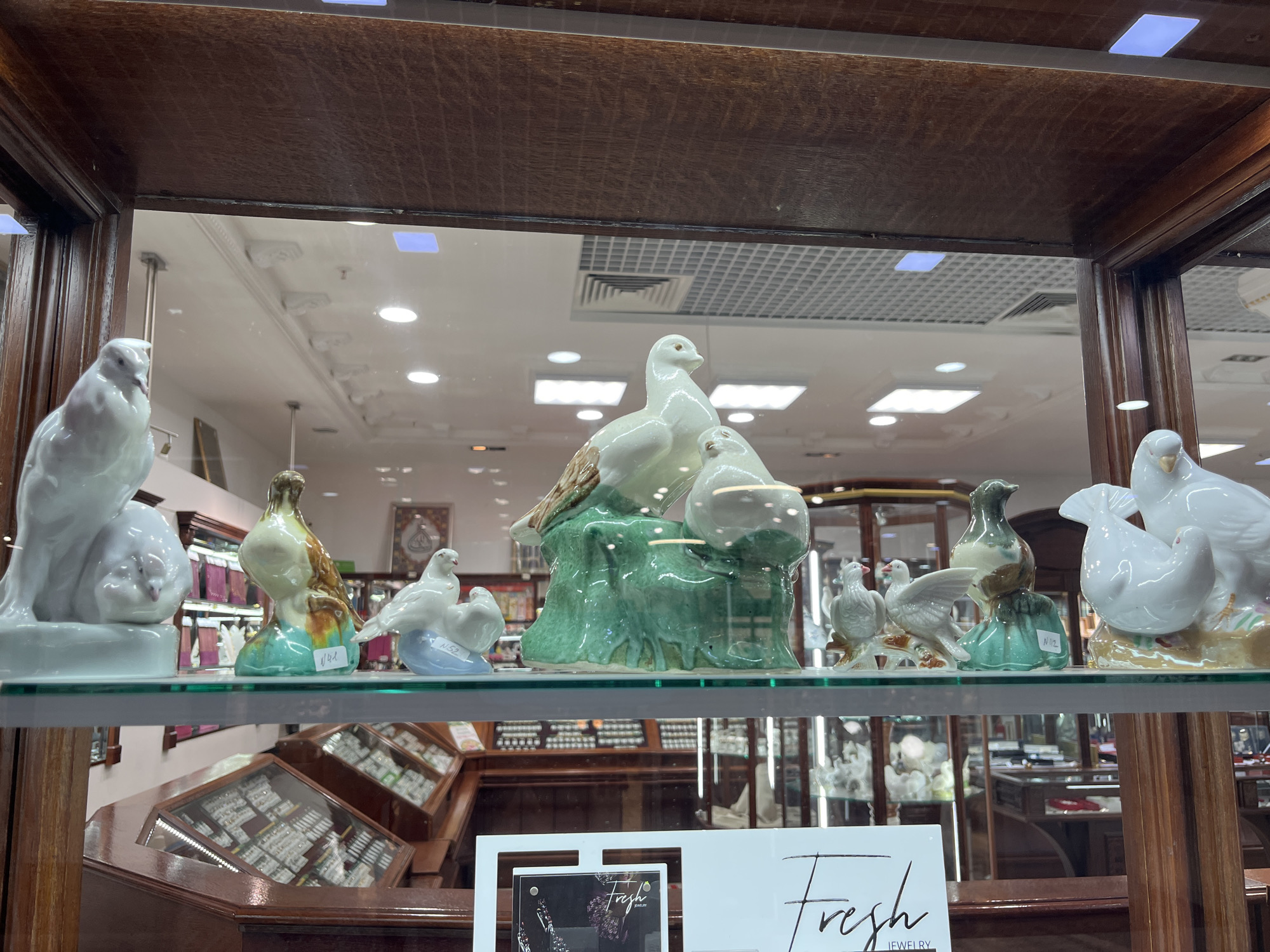 The image shows a shelf in a display case in a store, showcasing various porcelain bird figurines. The figurines are mostly white, with some featuring brown, green, or blue accents. They are arranged in a row on the glass shelf, with price tags visible on some of them. The background reveals more display cases and store merchandise. The store appears to be located in Izhevsk, Russia, as indicated by the "Fresh Jewelry" advertisement on the bottom shelf.