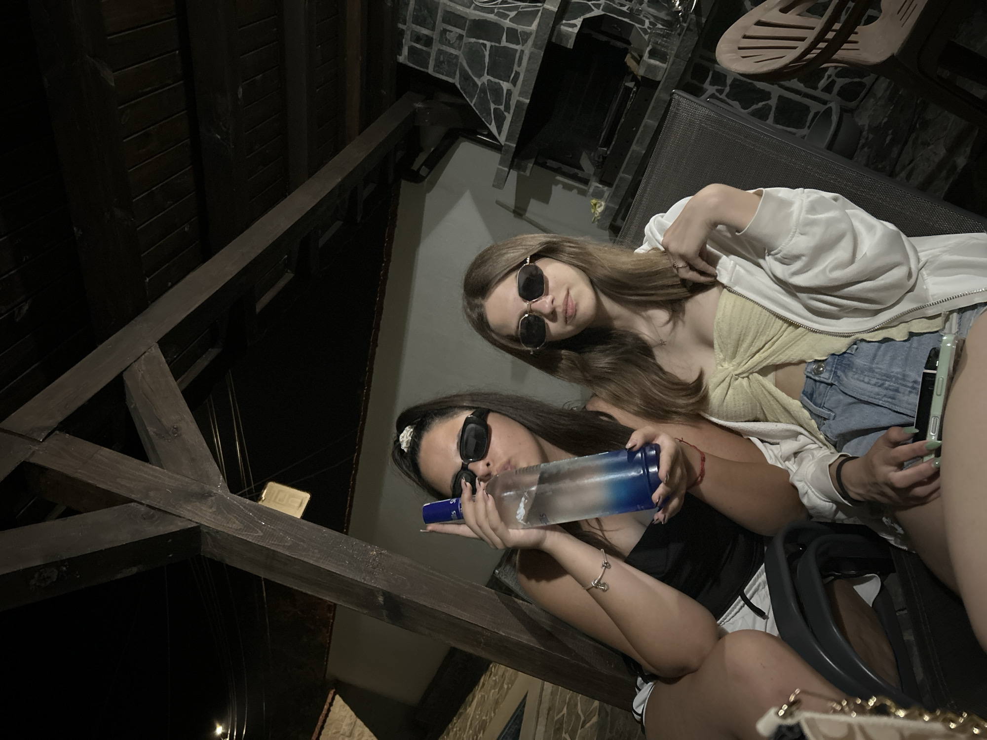 The image depicts two young women seated outdoors at night under a wooden pergola structure in the city of Unknown, Unknown. The scene is illuminated by ambient lighting, casting some areas into shadow.
The woman on the left has dark hair pulled back in a high ponytail secured with a white scrunchie. She wears dark, rectangular sunglasses and a black strapless or tank top. She is holding a clear glass bottle with a blue base and blue cap, consistent with a vodka bottle, to her mouth as if drinking. A silver charm bracelet is visible on her left wrist. She is seated in a dark chair, wearing white shorts with a dark trim.
The woman on the right has long, straight brown hair and wears light-colored, hexagonal-shaped sunglasses. She is dressed in a yellow twisted crop top, light-wash denim shorts, and a white zipped hoodie draped open over her shoulders. She holds a light green smartphone in her right hand, resting on her lap. A red string bracelet is visible on her left wrist. She is seated in a dark-colored mesh chair.
In the background, to the right, a large stone barbecue or outdoor fireplace is visible. Above, dark wooden beams and rafters form the overhead structure of the pergola. In the upper left background, a light source is visible, along with electrical wires. Further to the left, a light-colored building wall and the edge of a swimming pool are discernible. A light-colored handbag with a gold chain strap is partially visible in the bottom left corner of the frame. A small, light-colored plaque or sign is hanging from one of the wooden beams in the upper middle background.