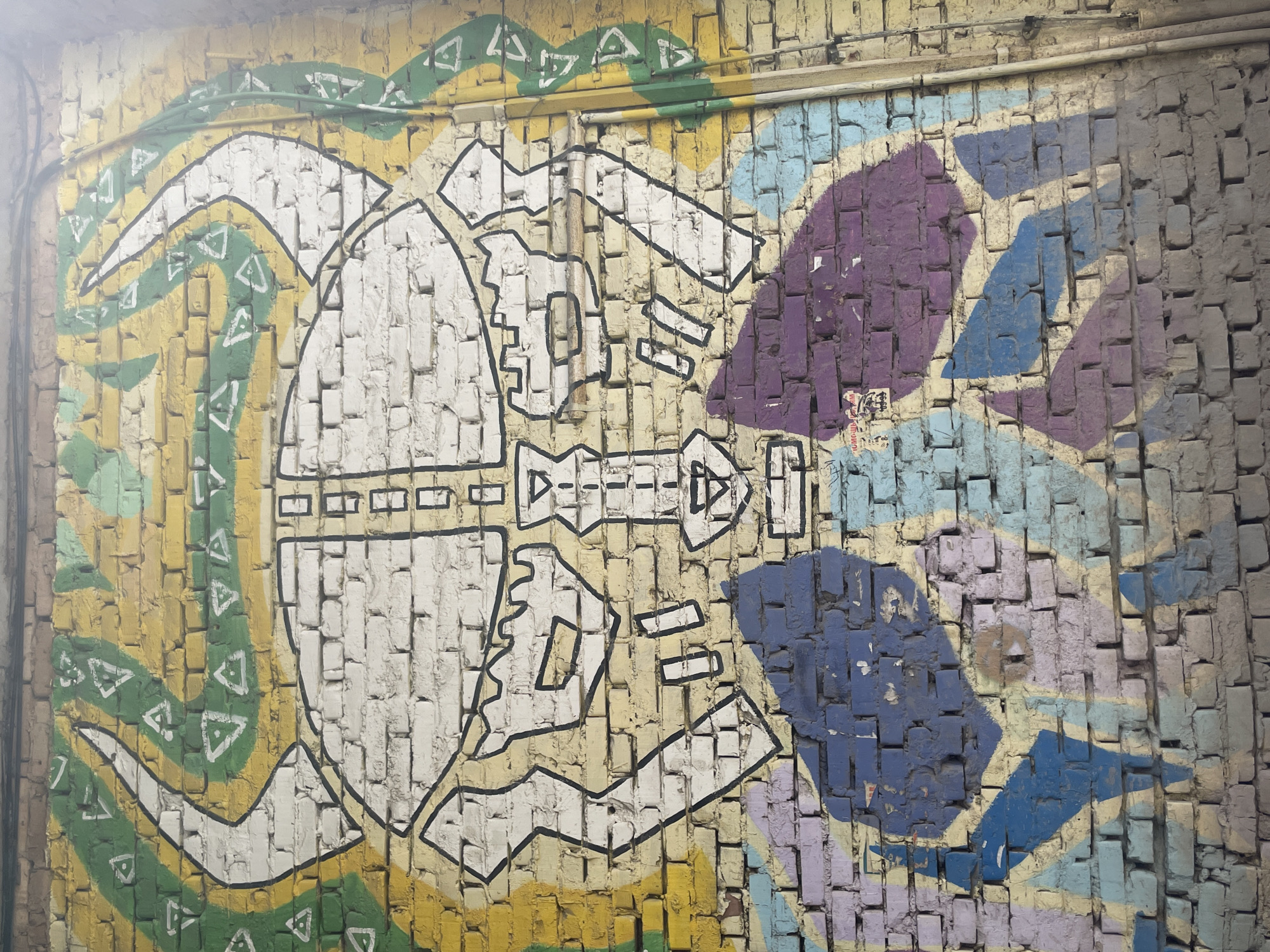 This is a photograph of graffiti on a brick wall in Izhevsk, Russia. The graffiti depicts a stylized, mask-like face with large horns extending upwards. The face is outlined in black and filled with white, with some abstract colorful shapes in blue and purple forming the lower part of the face or neck. Behind the face, there are swirling yellow and green patterns with white triangular shapes. The brick wall is weathered and shows signs of age. Two vertical pipes run along the right side of the wall. The overall scene suggests an urban outdoor environment.