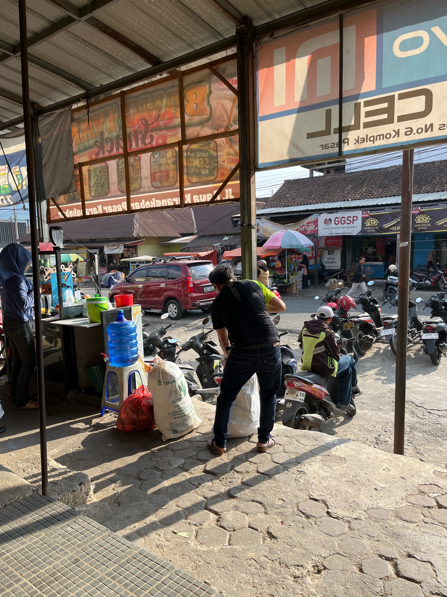 The image depicts an outdoor market scene in Karangkobar, Indonesia. In the foreground, a person wearing a blue hijab and dark pants stands at a food stall, while another person in a black t-shirt and jeans is bent over, possibly working. Several motorcycles are parked, and a red van is visible in the background. Various stalls are set up, displaying goods and advertising. The overall environment suggests a bustling marketplace with a mix of commercial activity and daily life.