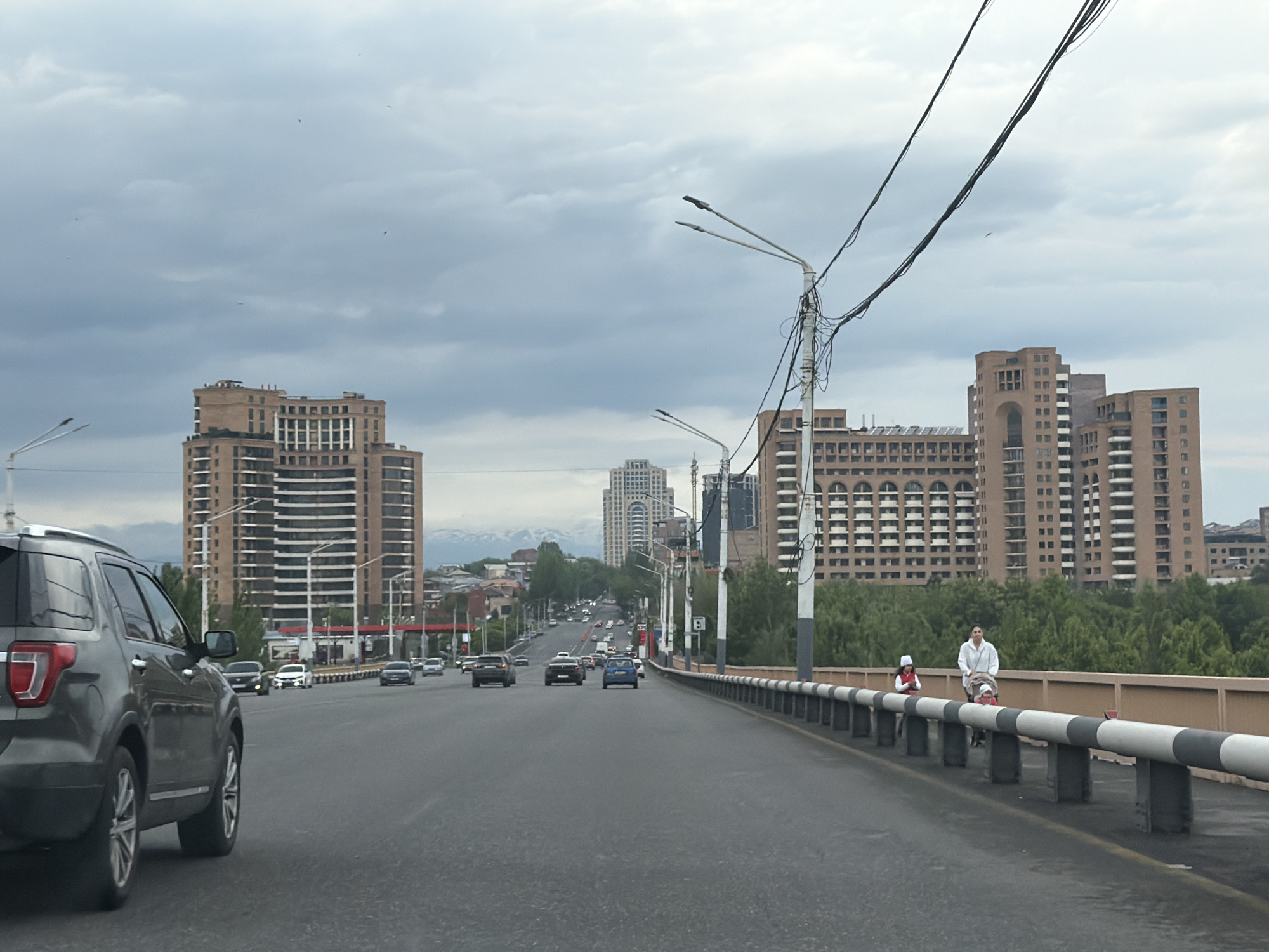 The image shows a wide street in Yerevan, Armenia. On the left, a dark gray Ford Explorer SUV is partially visible. The street stretches into the distance, with several other cars driving in the same direction. Tall, beige apartment buildings line both sides of the street. The buildings are built in a modern style with repetitive window patterns and arches. In the background, snow-capped mountains are faintly visible. On the right side of the street, a pedestrian walkway is lined with a black and white striped metal railing. A woman in a white coat and a child in a red hat are walking along the walkway, with the woman pushing a stroller. Above the street, power lines are suspended between lampposts. The sky is overcast with gray clouds.