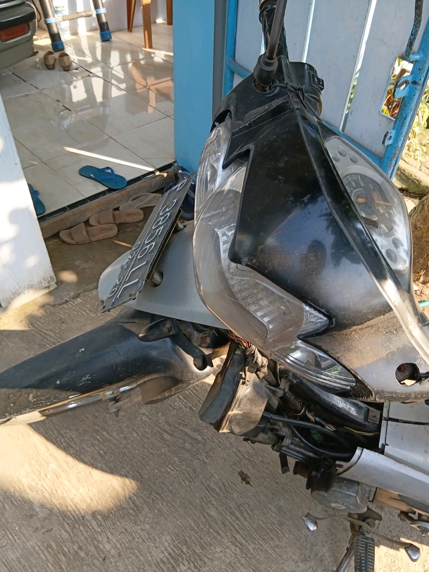 The image shows a motorcycle parked on a concrete surface outdoors, likely in Tanggul Wetan, Indonesia. The motorcycle is partially in shadow. The front of the motorcycle is visible, including the headlight assembly, front fender, and a license plate attached to the front. The license plate is dark colored with white characters. The motorcycle appears to be dusty. In the background, there is a tiled floor with several pairs of sandals and flip-flops scattered on it. A blue structure, possibly a fence or railing, is also visible. Sunlight is casting shadows on the ground, indicating it is daytime.