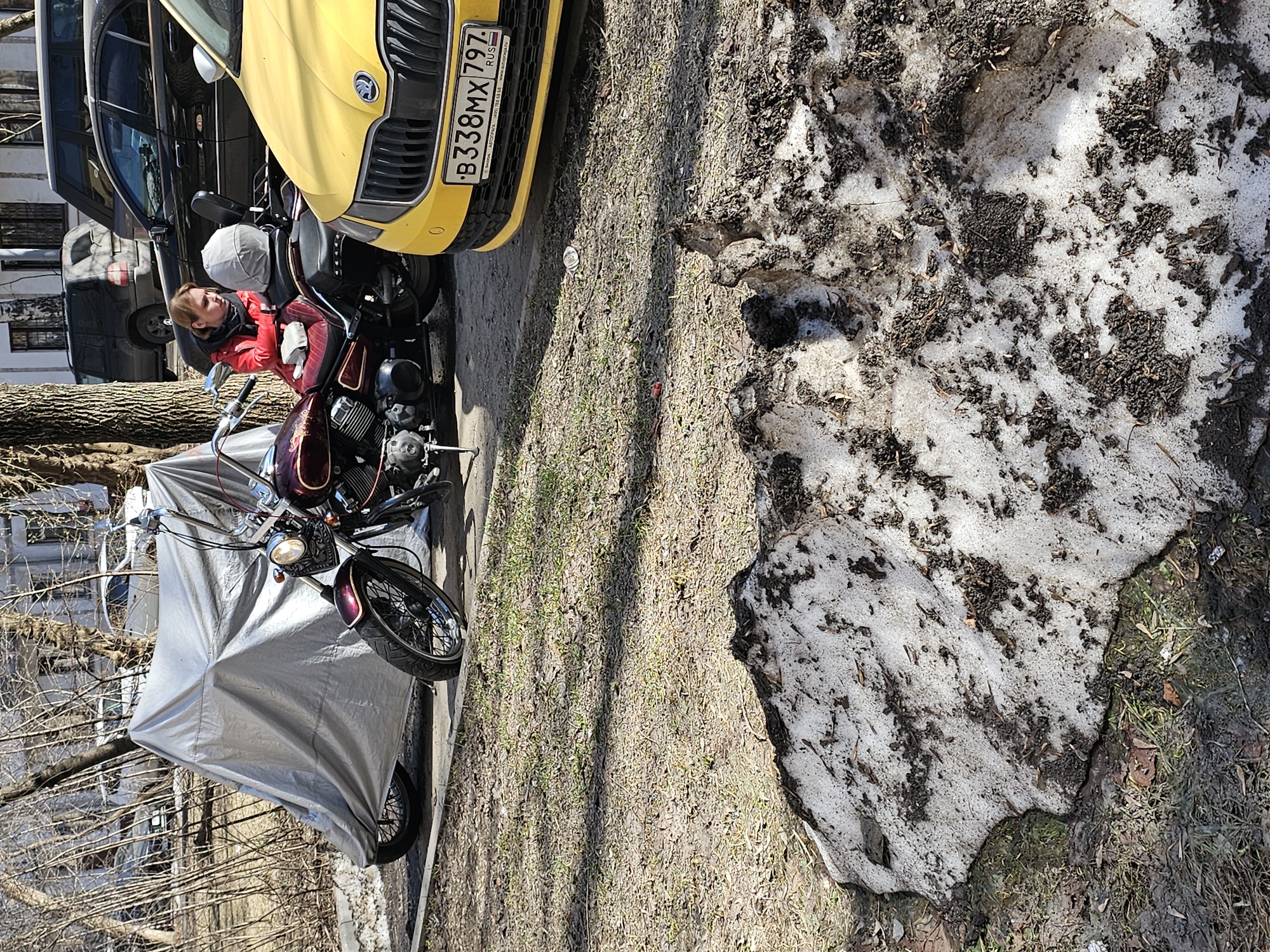 The image depicts a street scene in Khimki, Russia. A yellow Škoda car with license plate B338MX797 is parked next to a motorcycle partially covered by a silver tarp. A woman wearing a red jacket and a gray helmet sits on the motorcycle. Patches of melting snow are visible on the ground next to the motorcycle and car. Other vehicles are parked behind the Škoda. Bare trees suggest it is early spring. The ground is covered with grass and dirt.