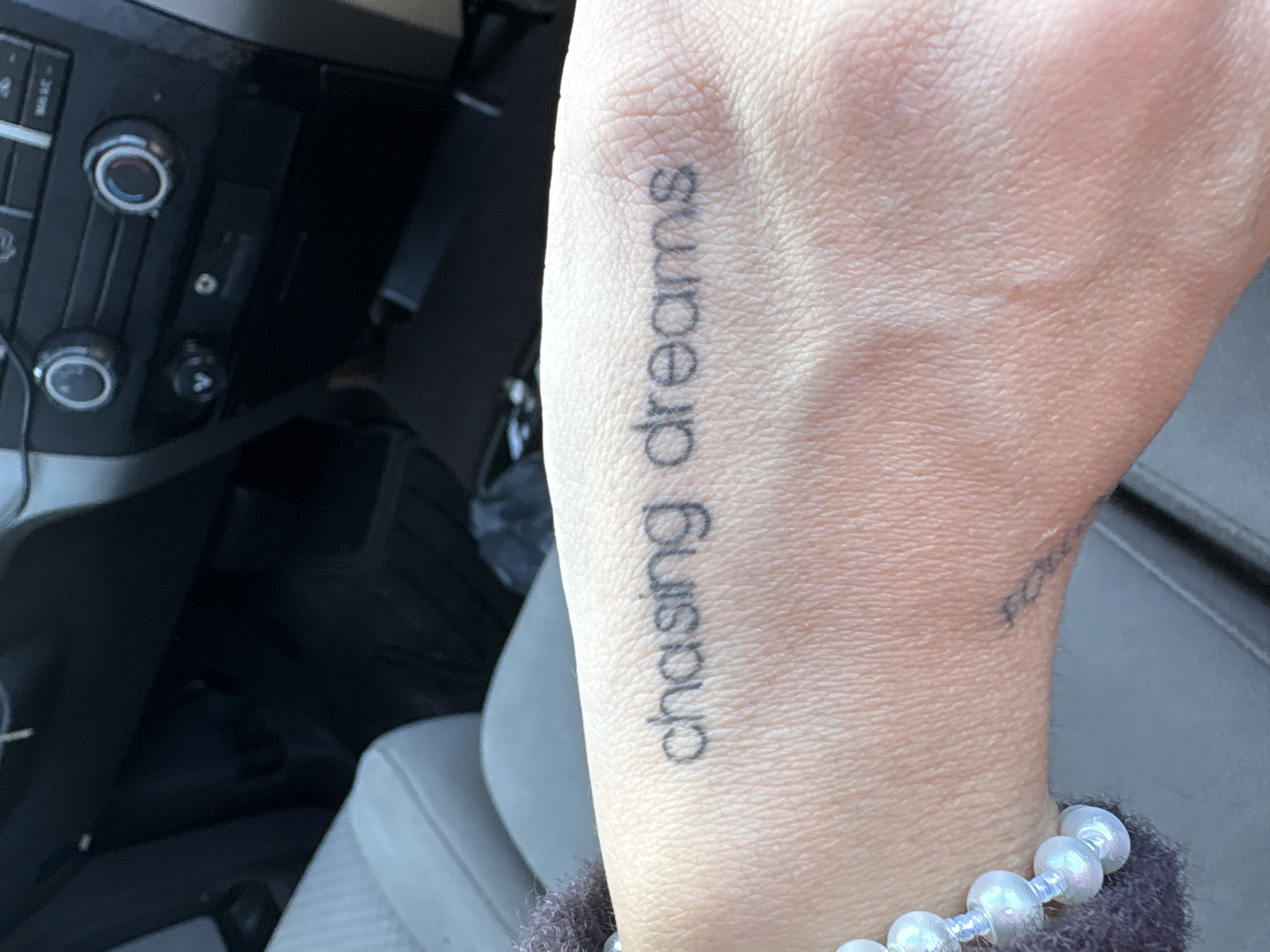 A person's hand is visible, featuring a tattoo that reads "chasing dreams" in a sans-serif font oriented vertically along the wrist and forearm. The hand also has a bracelet of iridescent white beads. The setting appears to be the interior of a vehicle, with a dashboard visible in the background. The dashboard includes climate control knobs and buttons. The seats are gray and appear to be upholstered in a cloth or synthetic material. The location is likely within Long Beach, California, USA as requested, but there are no visual cues to definitively identify a specific landmark or street within Long Beach.
