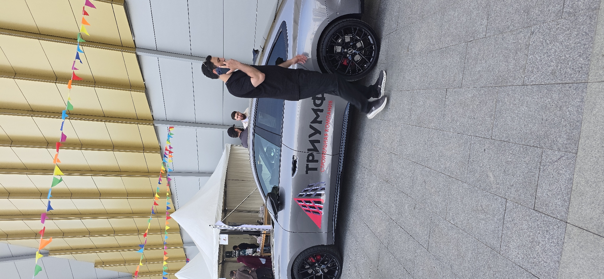 In Grozny, Russia, a grey car is parked on a paved surface. The car has "ТРИУМФ" and "СТРОИТЕЛЬНАЯ КОМПАНИЯ" printed on its side. A man in a black t-shirt and pants is walking past the car, holding a mobile phone to his ear. Behind the car, two white tents are set up, and several people are visible in the background. Colorful triangular flags are strung across the top of the image, against a building with a gold facade.
