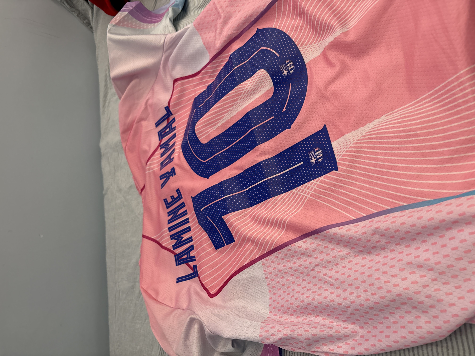 Here's a detailed description of the image:
**Scene Description:**
The image shows a pink sports jersey with the number "10" printed in large, blue font on the back. The name "LAMINE V YMAL" is printed vertically to the left of the number, also in blue. The background of the jersey features white lines radiating from the number, and a subtle pink dotted pattern in the lower right corner. The number "10" is also decorated with small images of the FC Barcelona team crest. The jersey appears to be lying on a grey surface.
**Location:**
The location is unknown. There are no clues in the image to identify the city or specific setting.