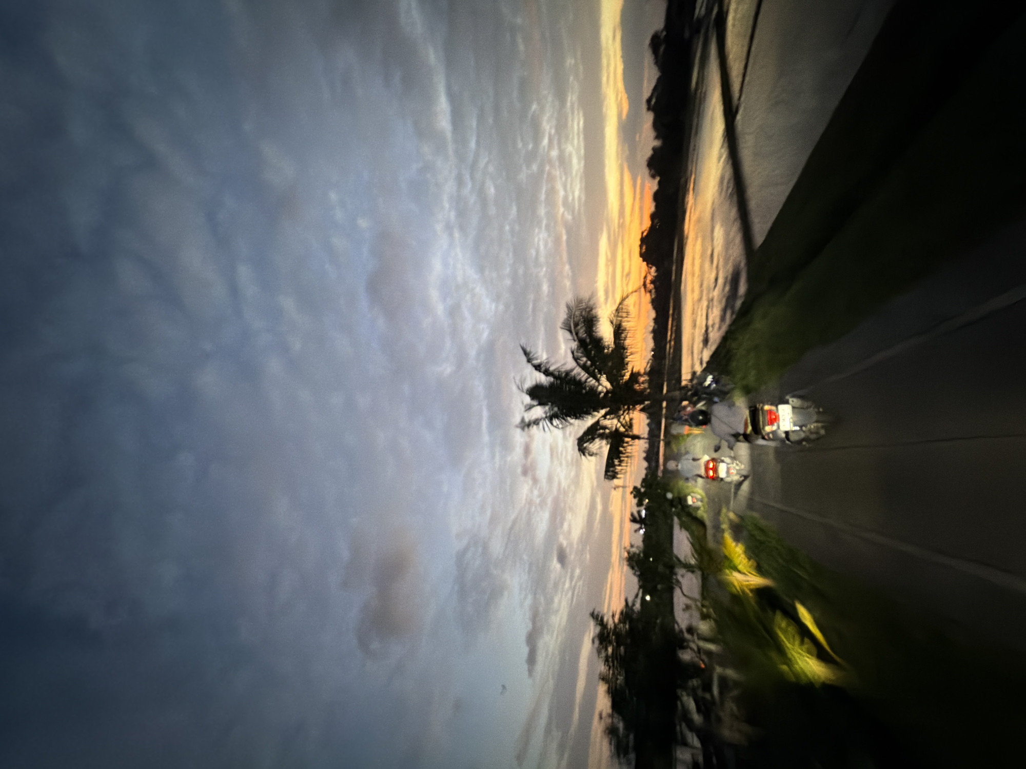 The image depicts a sunset scene on a road in North Kuta, Indonesia. A palm tree is visible on the side of the road. There are two motorcycles with riders visible in the distance, traveling along the road. The sky is filled with clouds, transitioning from darker blues on the left to orange and yellow hues near the horizon. The road surface is illuminated by headlights and appears wet. Vegetation lines the side of the road. A body of water is visible on the right side of the road, reflecting the colors of the sunset.