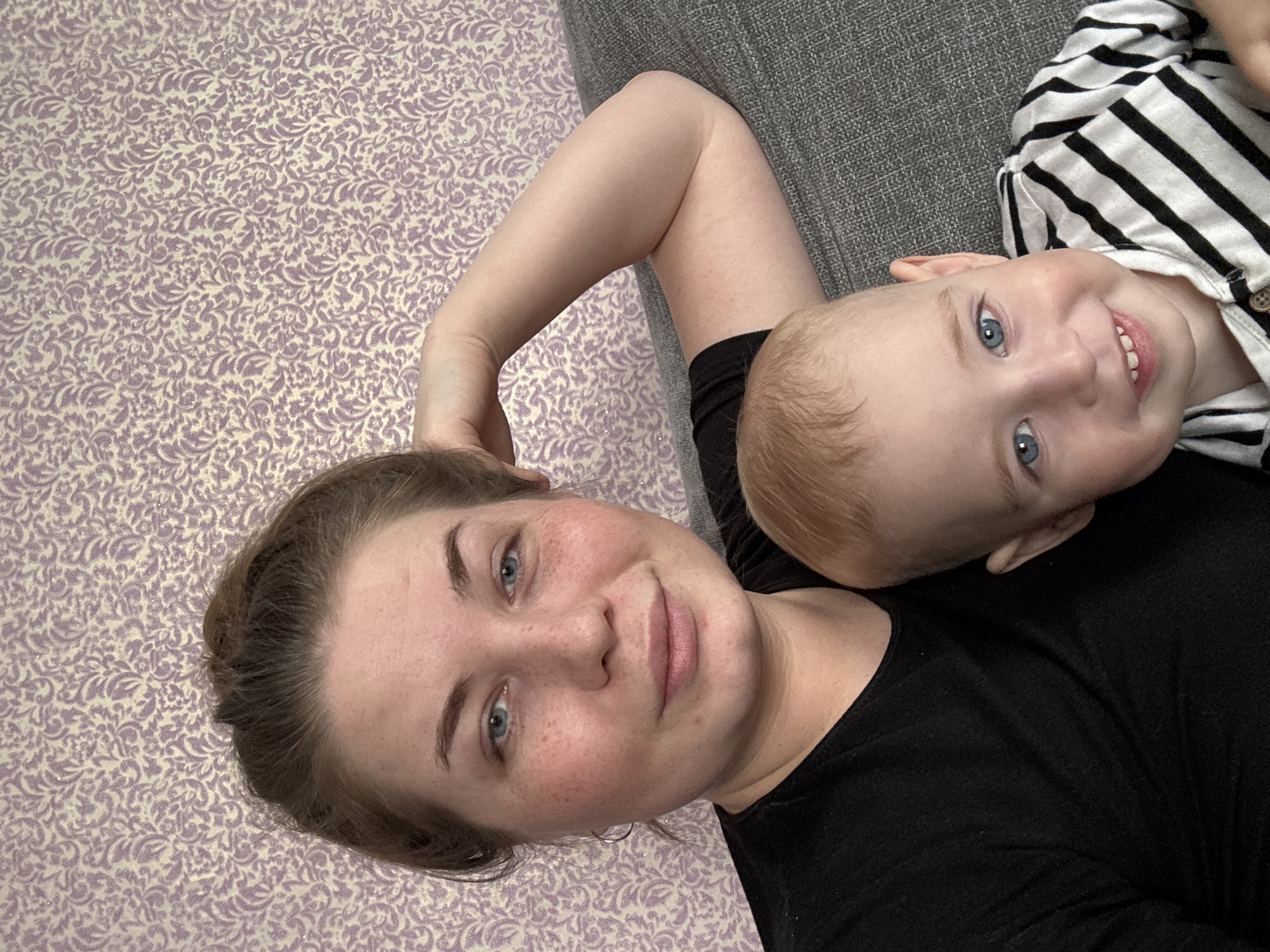 The image depicts a woman and a young child lying down together. The woman is on her back, wearing a black t-shirt. She has fair skin, light brown hair pulled back, and blue eyes. Her right arm is bent and resting above her head. The child is lying partially on the woman's chest. The child has fair skin, blonde hair, and blue eyes, and is wearing a black and white striped shirt. They are both smiling and looking at the camera. The background behind the woman is a patterned purple and white surface, possibly a wall or a piece of furniture. To the right of the woman is a gray surface, possibly a couch. There is no indication of the location being in Уральское, Russia, and the location cannot be determined from the image.