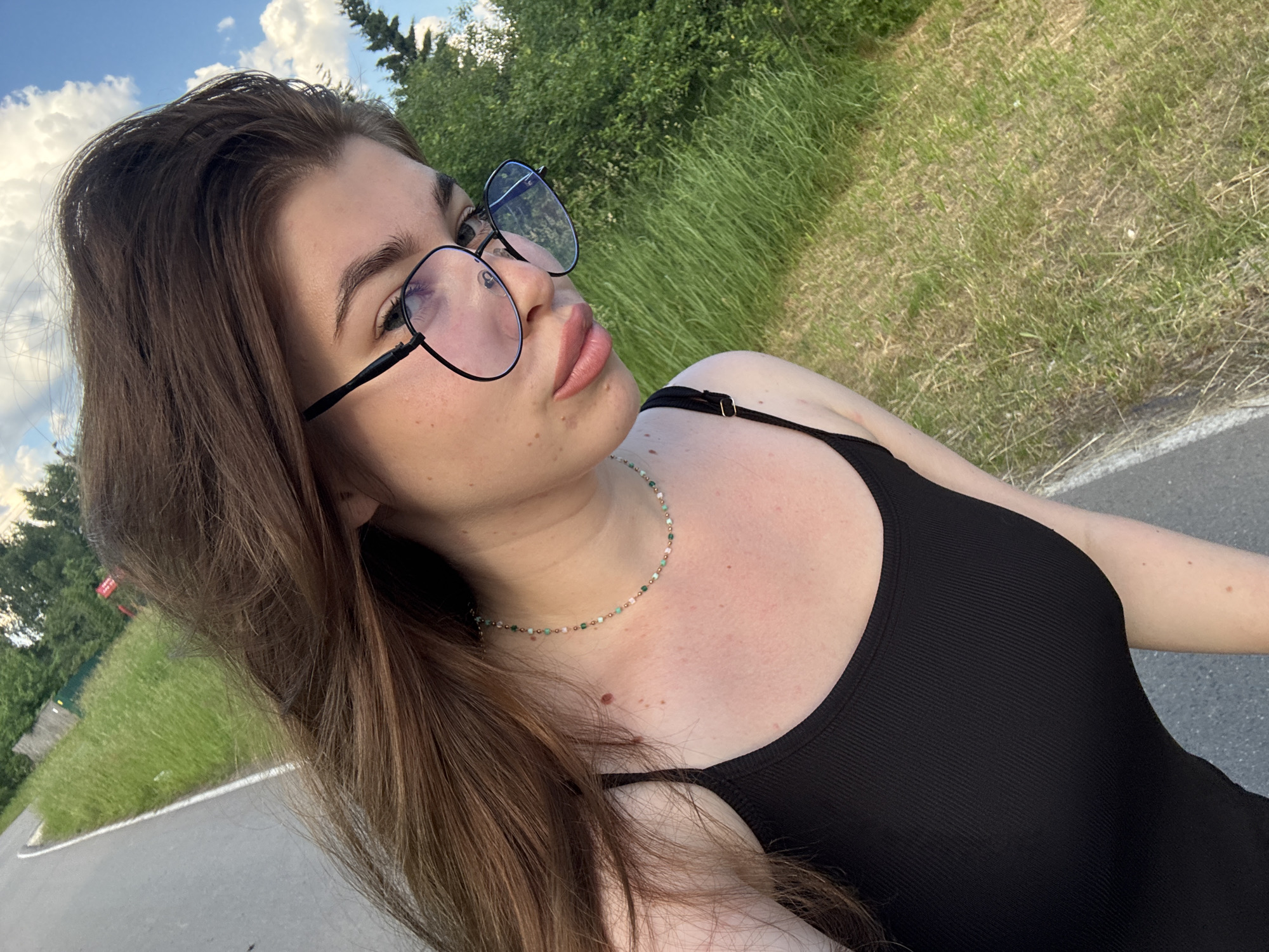 A young woman with long brown hair and glasses is posing for a selfie outdoors. She is wearing a black tank top and a beaded necklace. The background consists of a paved path, green grass, and trees. The sky is visible with some clouds. The overall setting appears to be a park or a recreational area.