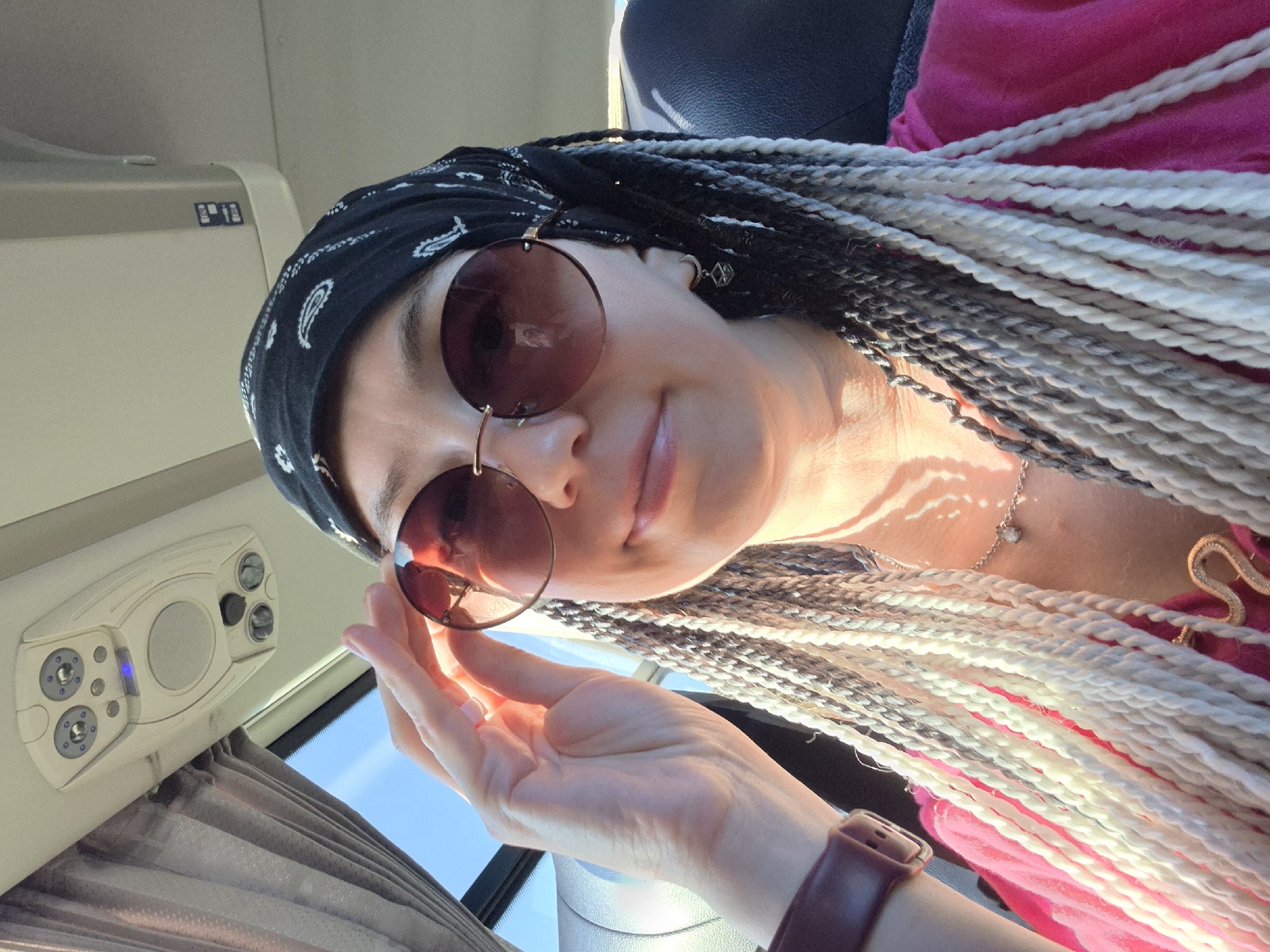 A woman with gray and white braided hair is wearing a black bandana and sunglasses, with her hand touching her sunglasses. She is wearing a pink shirt, a necklace, and an earring. The image appears to be taken inside a vehicle, possibly a bus, with a view of the sky and clouds through a window. The location in the city of Verkhniye Tatyshly, Russia is not discernible from the image itself.