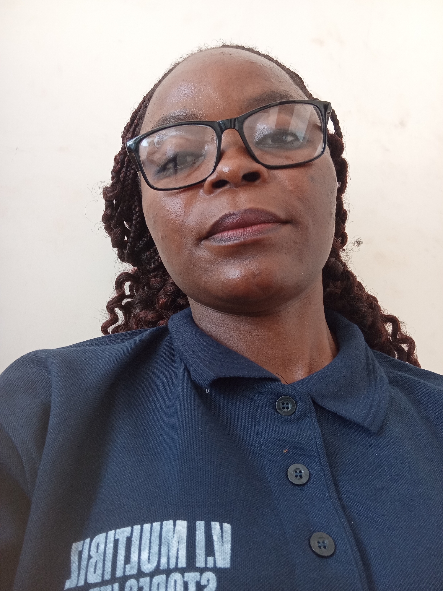 A medium shot shows a person wearing glasses and a dark blue collared shirt. The person has dark skin and braided hair. The background is a plain, light-colored wall. On the shirt, there is white text, but it is partially obscured and appears to be in a stylized font. The image is taken from a slightly low angle, looking up at the person. The lighting is even, and there are no shadows. The overall impression is that of a casual selfie. The image does not contain any specific landmarks or contextual clues that would allow for its location to be identified as Jalingo, Nigeria.