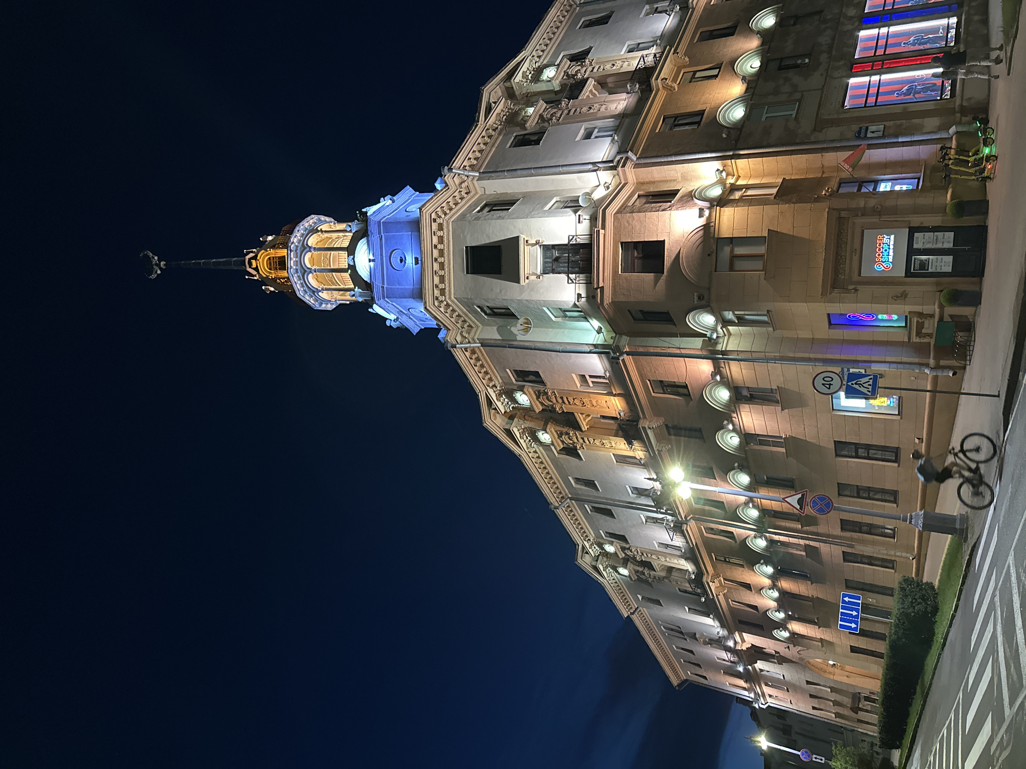The image shows a building in the city of Minsk, Belarus, at night. The building is illuminated, with a prominent tower topped with a spire. The tower is lit in blue and yellow, with ornate architectural details. The main body of the building is also lit with warm yellow and white lights, highlighting its windows and decorative elements. In the foreground, a street with crosswalk markings is visible. A person is riding a bicycle across the crosswalk, captured in motion blur. There are traffic signs and streetlights along the sidewalk. A shop named "SOCCER SHOP.BY" is visible on the ground floor, with illuminated windows displaying sports-related imagery. Several electric scooters are parked in front of the shop. The sky is dark blue, indicating nighttime.