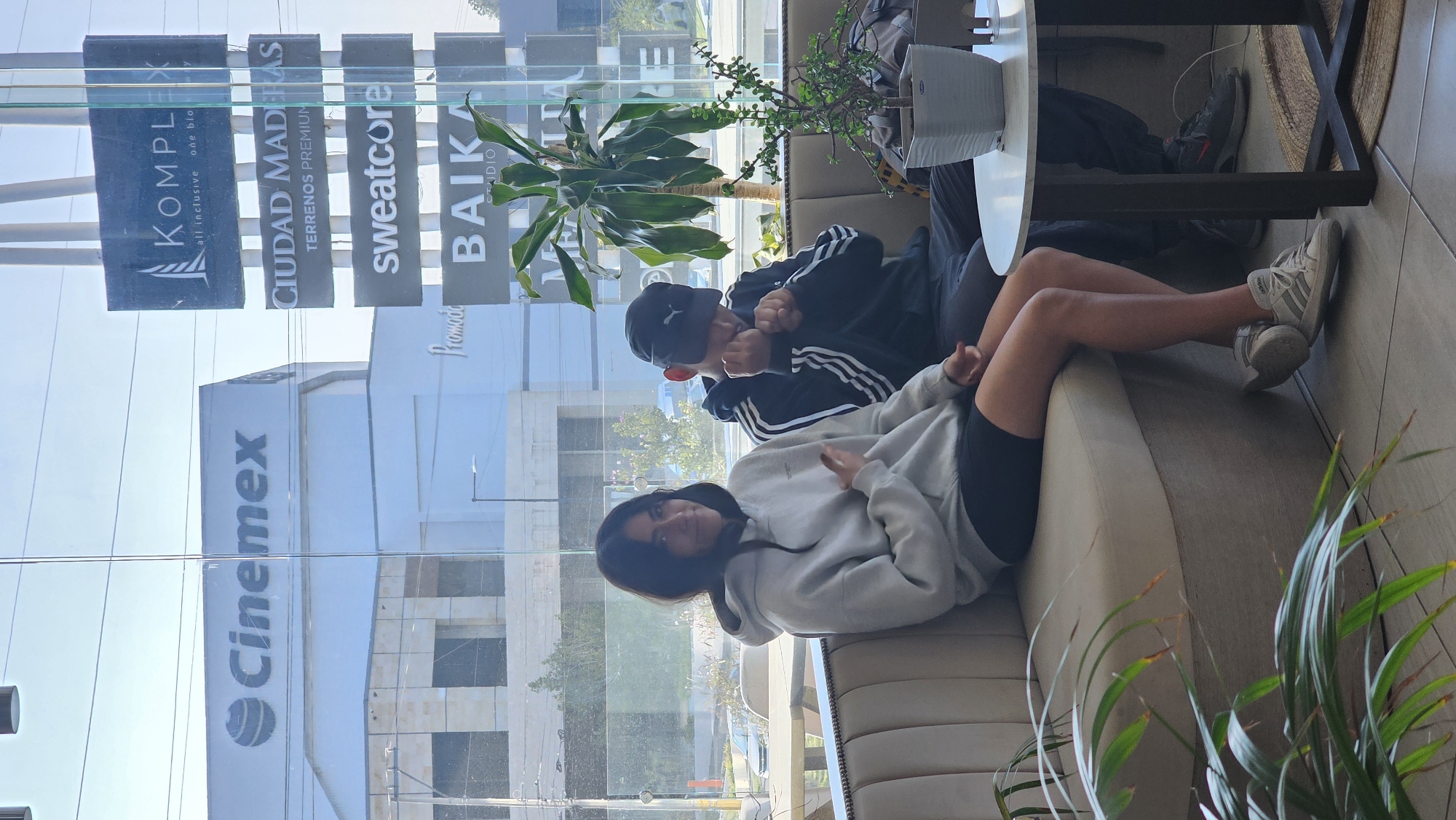 Here's a detailed description of the image:
**Scene:** The photo appears to be taken inside a commercial establishment, likely a cafe or waiting area, with a large window providing a view of a busy street and buildings in the city of Puebla, Mexico.
**People:** Two young individuals are seen lounging on a cushioned sofa. One, wearing a light-colored hoodie and dark shorts, is closer to the camera and looks towards it with a casual pose. The other person, dressed in a dark jacket with white stripes and a baseball cap, is partially obscured and facing away. The person facing away seems to be taking or faking a photo.
**Objects:**
* Sofa: A light-colored, cushioned sofa.
* Table: A round, white table.
* Plants: Various potted plants are placed around the seating area, both on the table and near the sofa.
* Buildings: Visible through the window are commercial buildings, showcasing signs like "Cinemex" (a cinema chain), "KOMPLEX", "CIUDAD MADERAS", "sweatcore", and "BAIKA".
**Actions/Interactions:** The two individuals are relaxing and interacting with each other. The one closer to the camera is posing, while the other seems to be taking a photo.
**Environment:** The setting seems urban, with a mix of modern architecture and commercial activity. The presence of numerous advertisement signs suggests a bustling area.
**Location Clues:** The "Cinemex" sign, along with other brand names, might aid in pinpointing the exact location within Puebla. The presence of the multiple ad signs indicate a shopping center.
