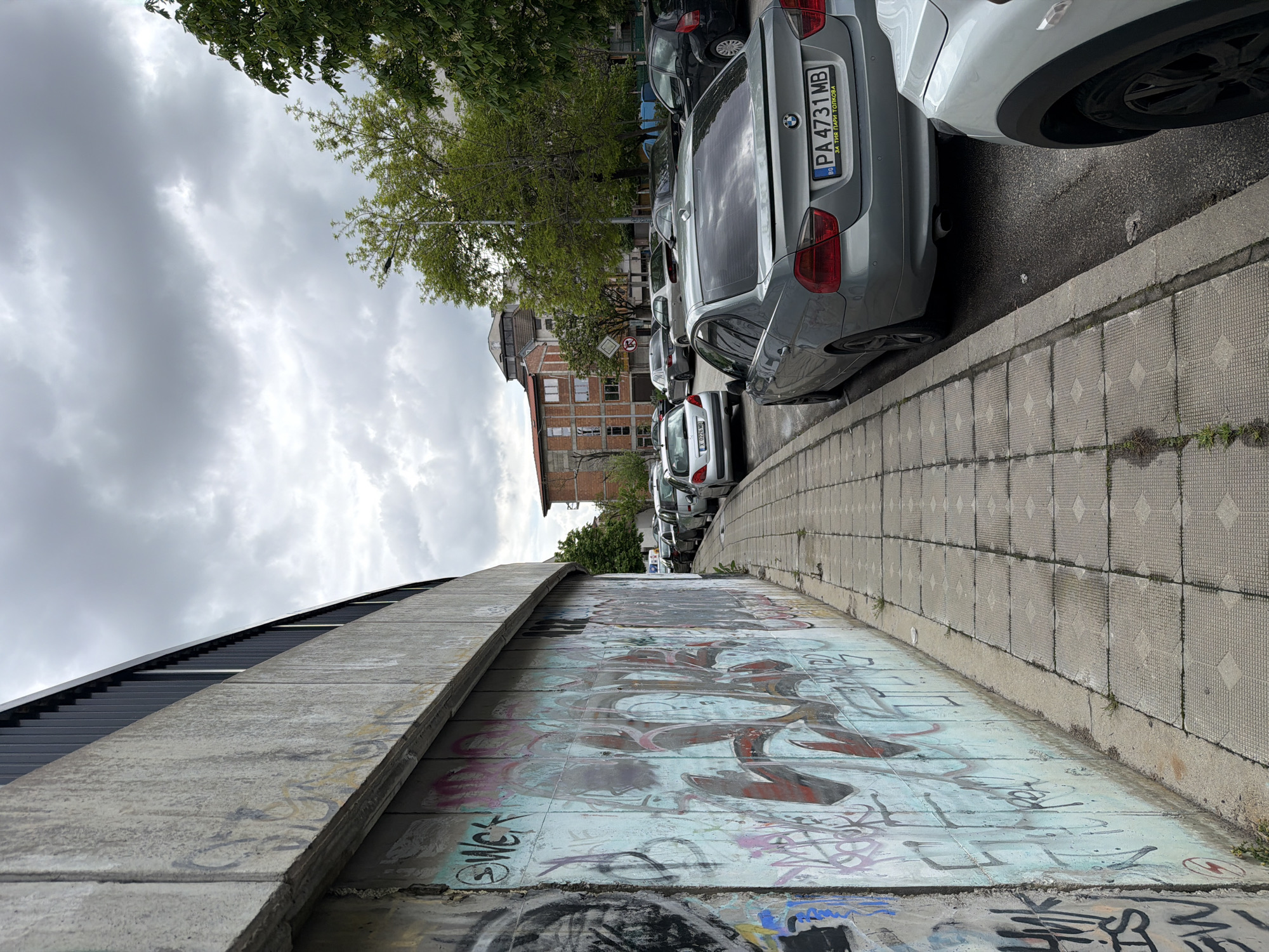 A sidewalk in Plovdiv, Bulgaria, is located next to a concrete wall covered in graffiti. A line of parked cars, including a gray BMW with license plate "PA 4731 MB," runs parallel to the sidewalk. In the background, there is a brick building. The sky is overcast with gray clouds, and trees are visible above the cars.
