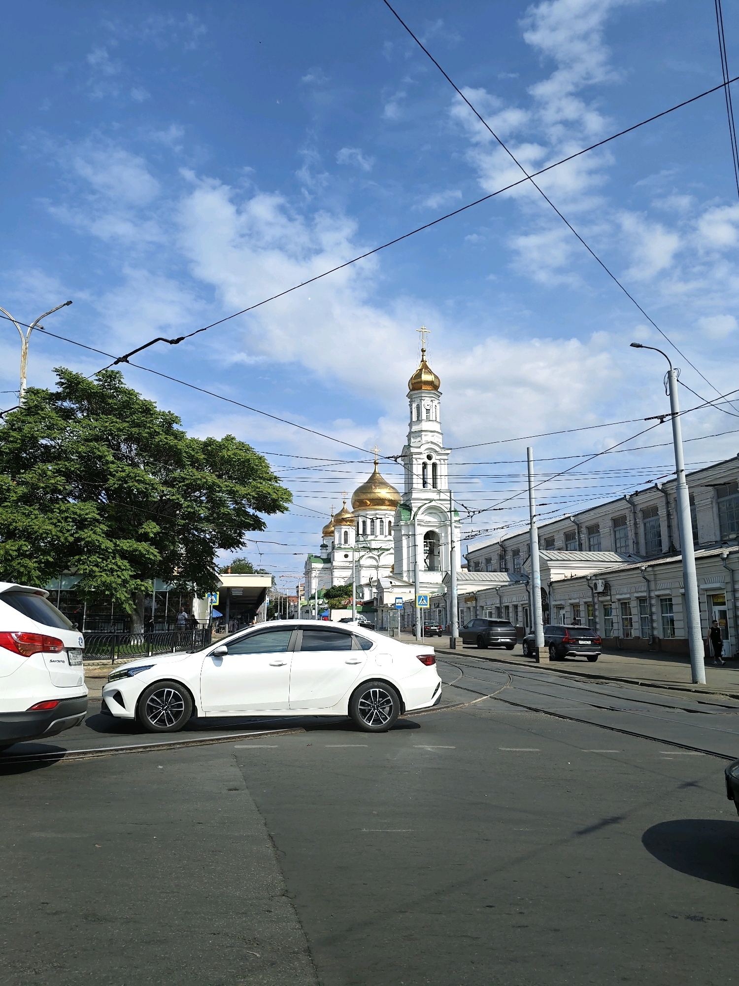 The image depicts a street scene in Rostov-on-Don, Russia, under a bright, cloudy sky. A white sedan is prominently featured in the foreground, positioned on an asphalt road with tram tracks. To the left, the rear of a white SUV is visible.
In the mid-ground, a large green tree stands to the left, and a building with a glass facade is behind it. Across the street, a white church with golden domes and a bell tower dominates the skyline, a clear landmark of religious significance in the city. Adjacent to the church are several multi-story buildings with a classical architectural style, featuring rows of windows and light-colored facades.
Several other vehicles, including dark-colored sedans and SUVs, are present on the road, some parked and others in motion. Power lines crisscross the sky, indicating the presence of urban infrastructure. A lamppost is visible on the right side of the image. Two individuals are seen walking on the sidewalk near the buildings on the right. The overall impression is that of a typical urban environment on a sunny day, with a blend of modern and historical architecture.