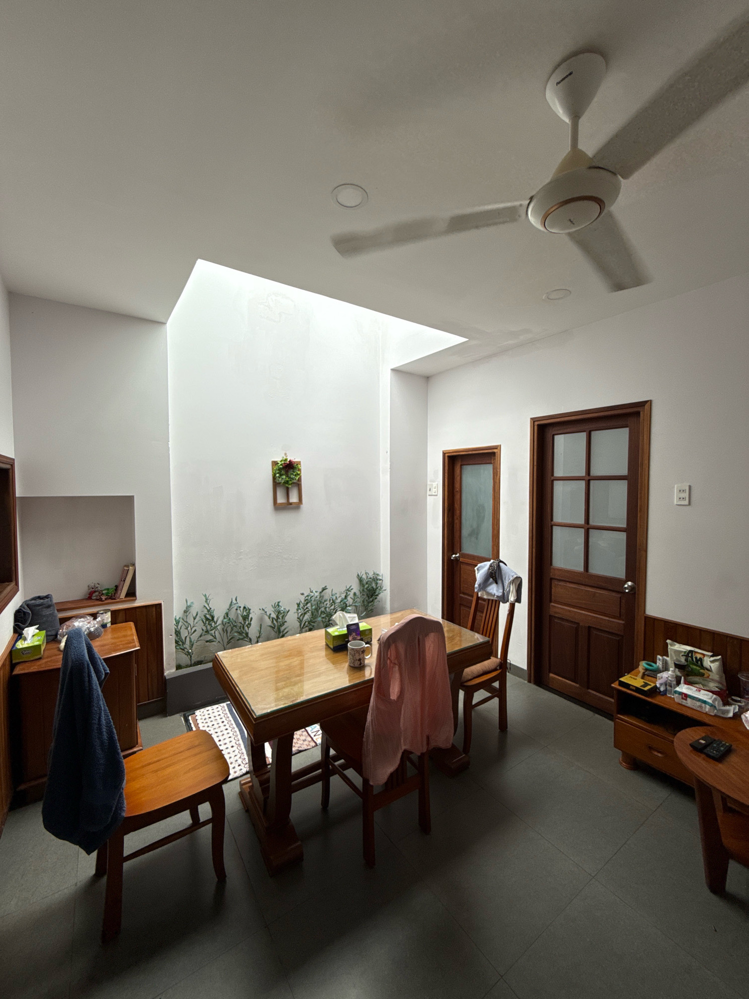 This is a wide-angle, eye-level shot of a dining area within a residential setting in Nha Trang, Vietnam. The room features a large wooden dining table with four wooden chairs. A dark blue towel hangs from the back of one chair, and a light pink garment is draped over another. Two wooden doors with glass panels are visible on the right side of the frame. The walls are painted white, with a skylight in the ceiling providing natural light. Decorative green foliage is painted along the lower portion of one wall. A small wooden cabinet with shelves is positioned to the left of the dining table, holding various items including tissue boxes and books. On the right, a wooden console table displays a variety of items, including tissue boxes, packaged goods, and a mug on the dining table. A ceiling fan is visible in the upper portion of the frame. The floor is tiled in a dark gray color. The overall scene suggests a casual and lived-in dining space.