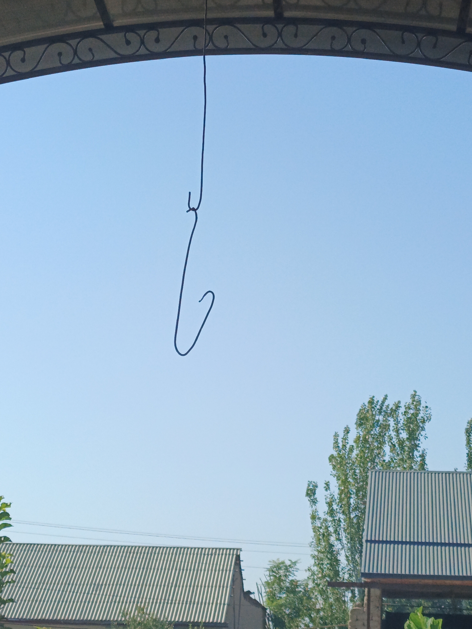 The image shows a blue sky with a dark wire hanging from an overhead structure. The wire is bent into a shape resembling a hook. Below the sky, there are parts of two buildings with corrugated metal roofs, one predominantly green and white striped, and another darker, possibly gray. Several tall trees with green foliage are visible in the background, along with some power lines. The overall scene appears to be an outdoor setting in a residential or industrial area. The image is taken from a low angle, looking upwards. The location is Jizzakh, Uzbekistan.