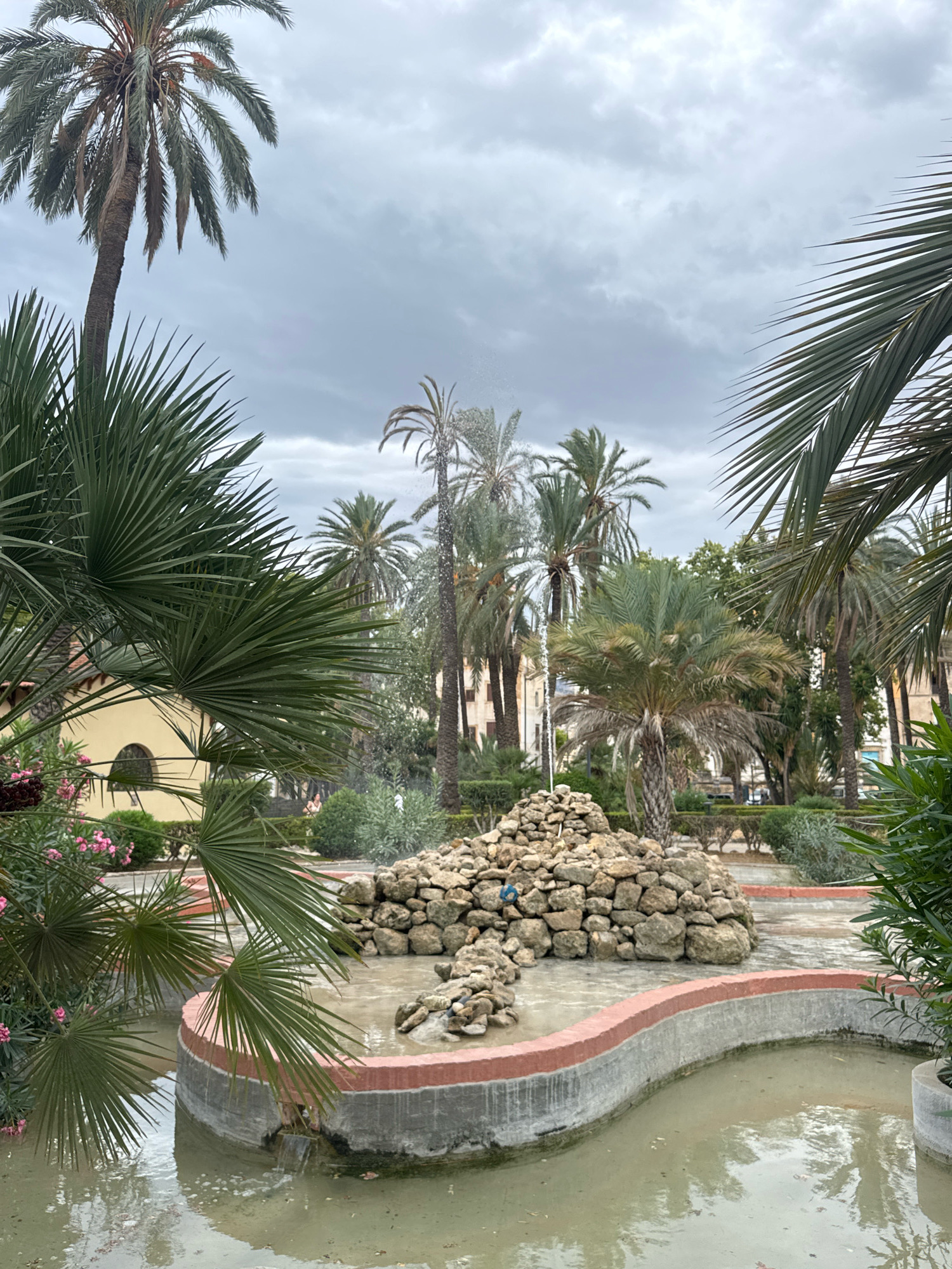 The image depicts a view of a dried-out, stone-filled fountain and surrounding garden area, likely in Palermo, Italy. The fountain, with its curved concrete basin lined with red trim, is mostly filled with stagnant, murky water and debris. A small mound of rocks sits in the center, suggesting it was once part of the fountain's decorative elements. Lush palm trees, some with long, drooping fronds, dominate the scene, with their leaves creating a canopy over the area. In the background, buildings with arched windows and other greenery are visible. The sky is overcast and gray, suggesting an impending or recent rainfall. The scene appears to be a public park or garden, possibly in a residential area, given the presence of buildings. The overall impression is one of disuse or neglect, with the fountain clearly not in active operation.