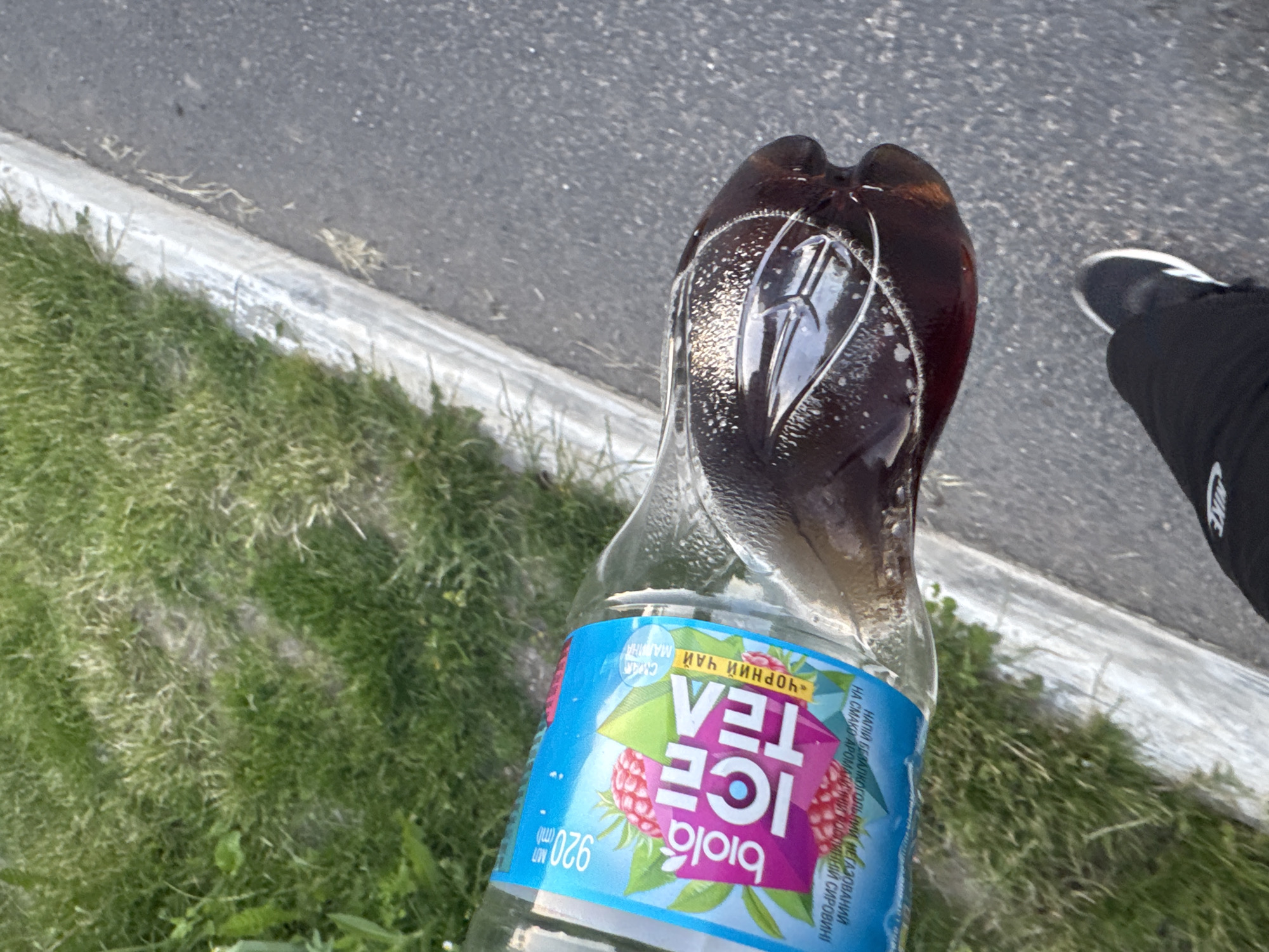 A transparent plastic bottle of Biola Ice Tea, with a raspberry and green geometric design on its label, is tilted to the right, with dark brown liquid filling about a third of its volume. The bottle rests on a patch of green grass next to a concrete curb, which separates the grass from a grey asphalt surface. A person's legs, clad in black pants and white-tipped sneakers, are visible in the lower right portion of the frame, suggesting they are standing on the asphalt. The scene is outdoors in daylight, with the lighting indicating it could be morning or late afternoon.