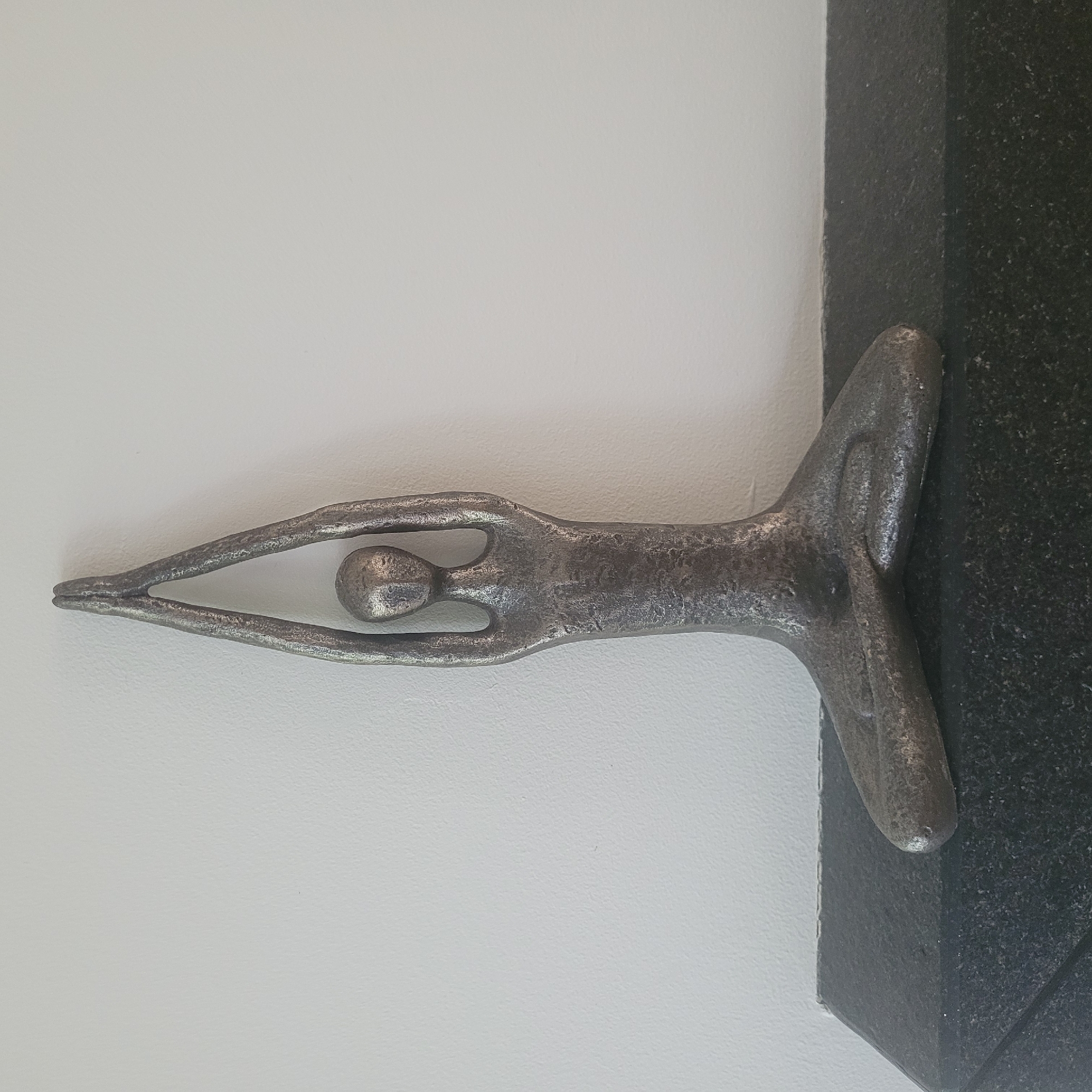 A metallic sculpture of a person in a meditative pose, with arms raised and hands clasped above the head, is positioned on a dark, speckled surface. The sculpture is silver-toned and appears to be made of a rough-textured metal. The background is a plain white wall with a slight texture. The surface the sculpture rests on appears to be a countertop or shelf, possibly made of granite. The image is a straightforward, frontal view of the sculpture. No specific location in Jūrmala, Latvia, can be identified from the visual information provided.